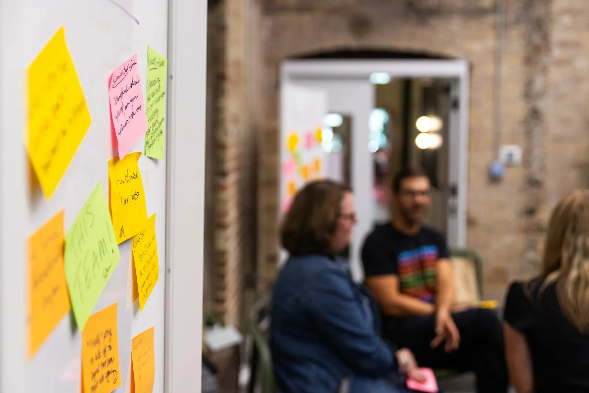 A wall with colorful sticky notes on a whiteboard or wall, with a blurred group of people sitting and talking in the background.