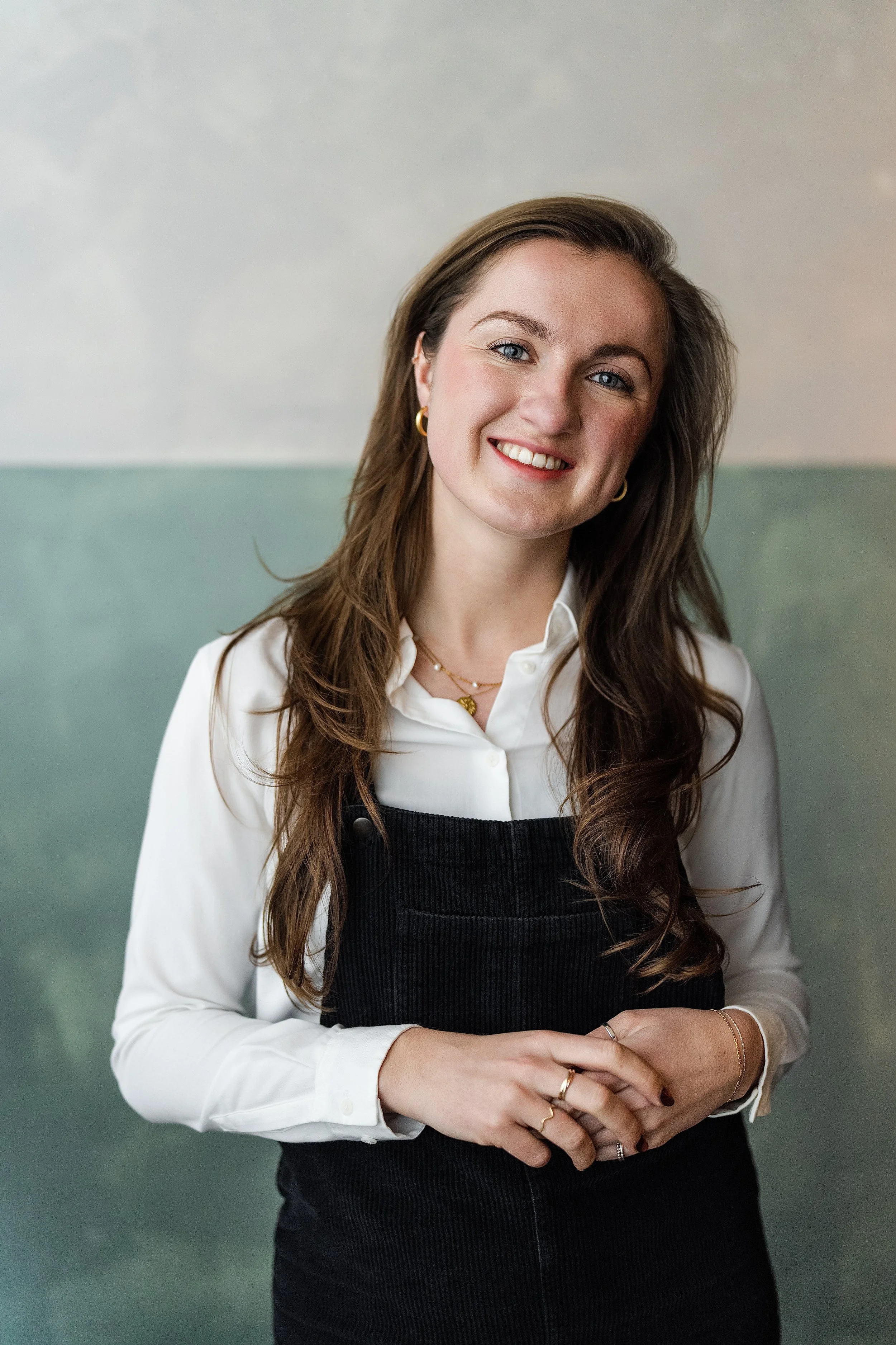 A smiling woman with long brown hair, blue eyes, wearing a white shirt and black overalls, standing against a light green and gray background.
Claire Shaw