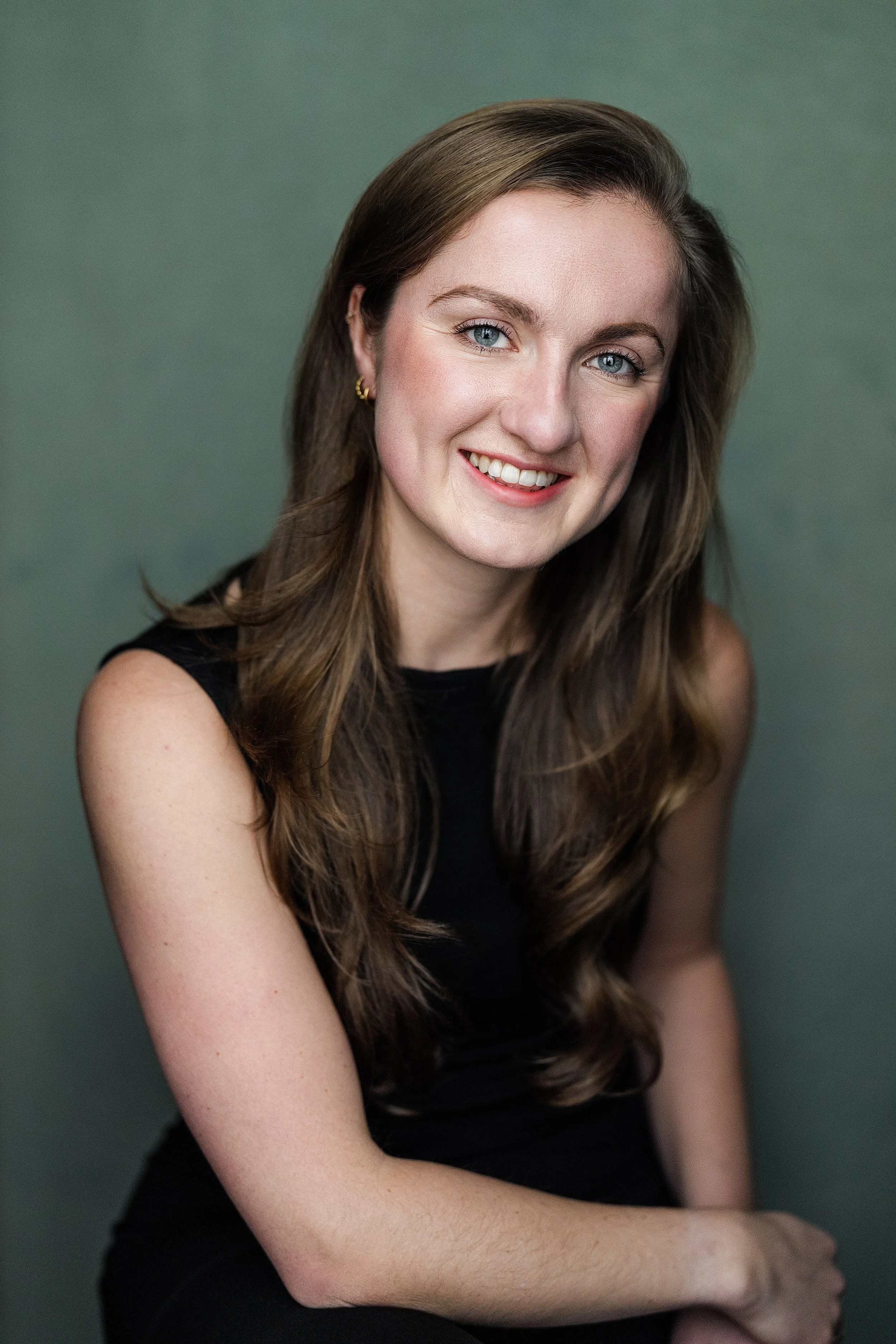 A young woman with long brown hair and blue eyes, smiling and wearing a black sleeveless top, sitting against a green background.
Claire Shaw