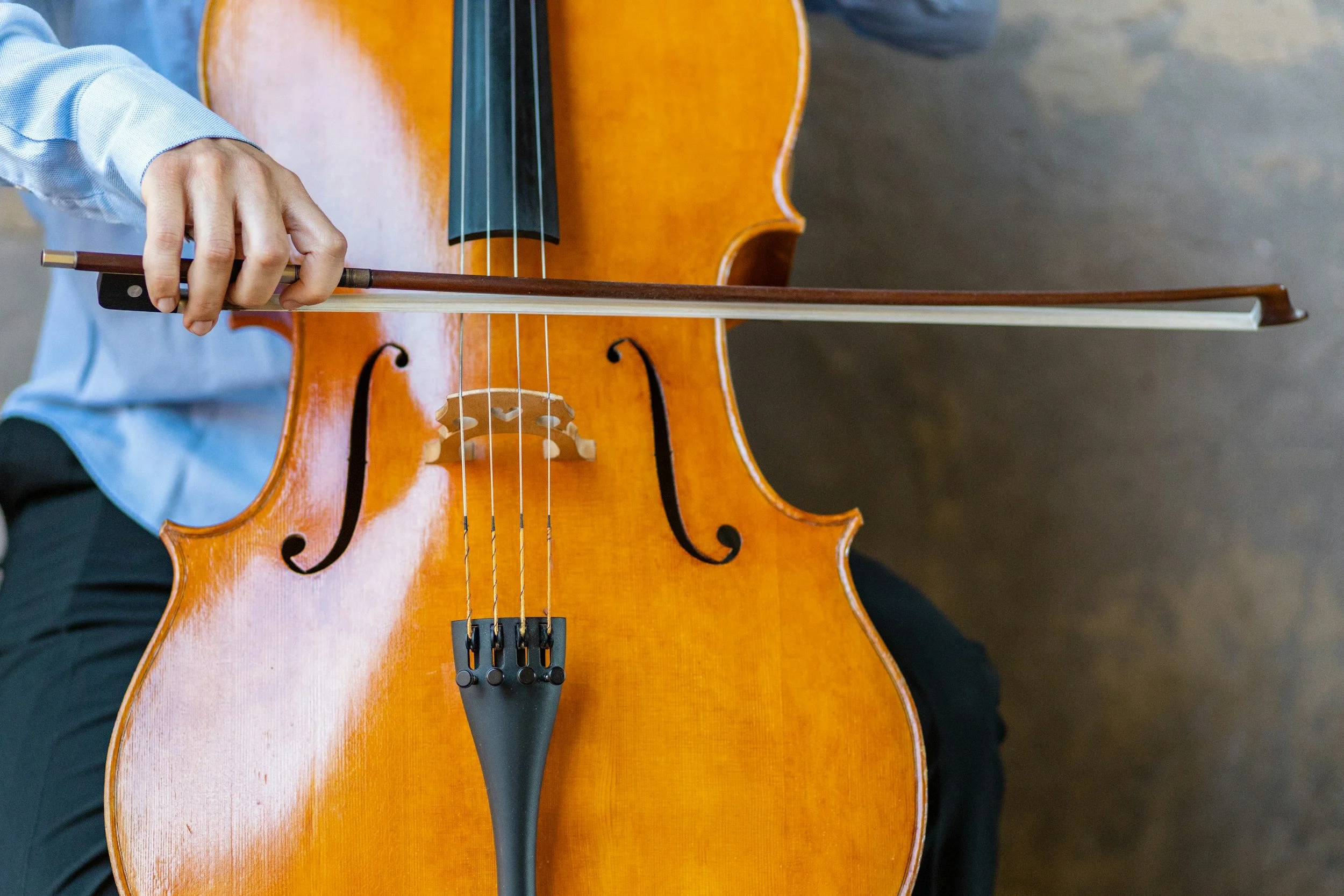Close-up of a person playing a wooden cello, focusing on their hand holding the bow and part of the cello's body.