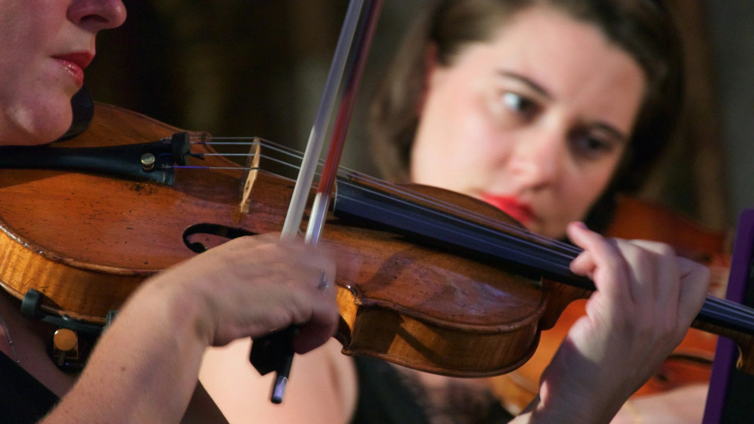 Close-up of a woman playing the violin with a bow, with another woman in the background watching her.