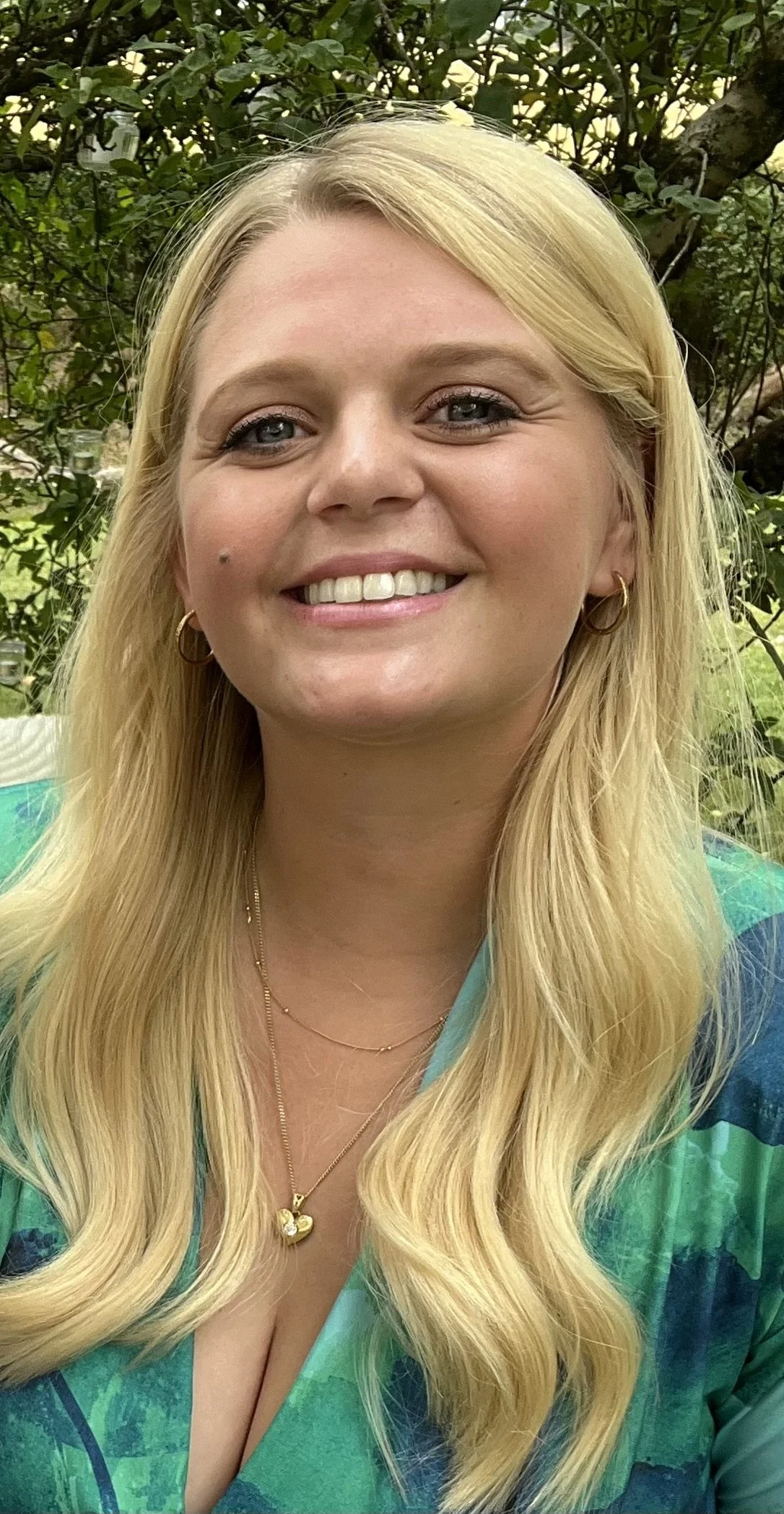 A woman with long blonde hair wearing a green patterned top and gold jewelry, smiling outdoors with greenery in the background.