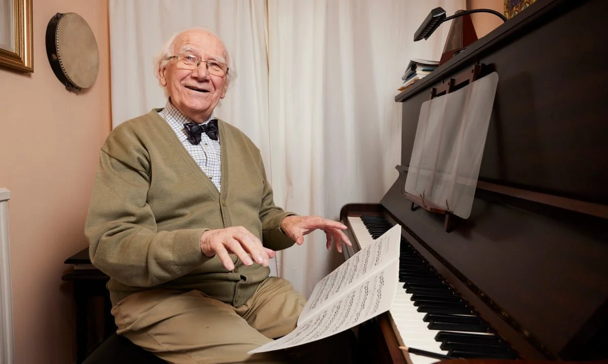 An elderly man smiling while sitting at a piano, holding sheet music in his hand.