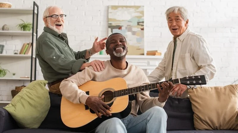 Three men enjoying music together in a living room, one playing a guitar and the other two smiling and laughing.