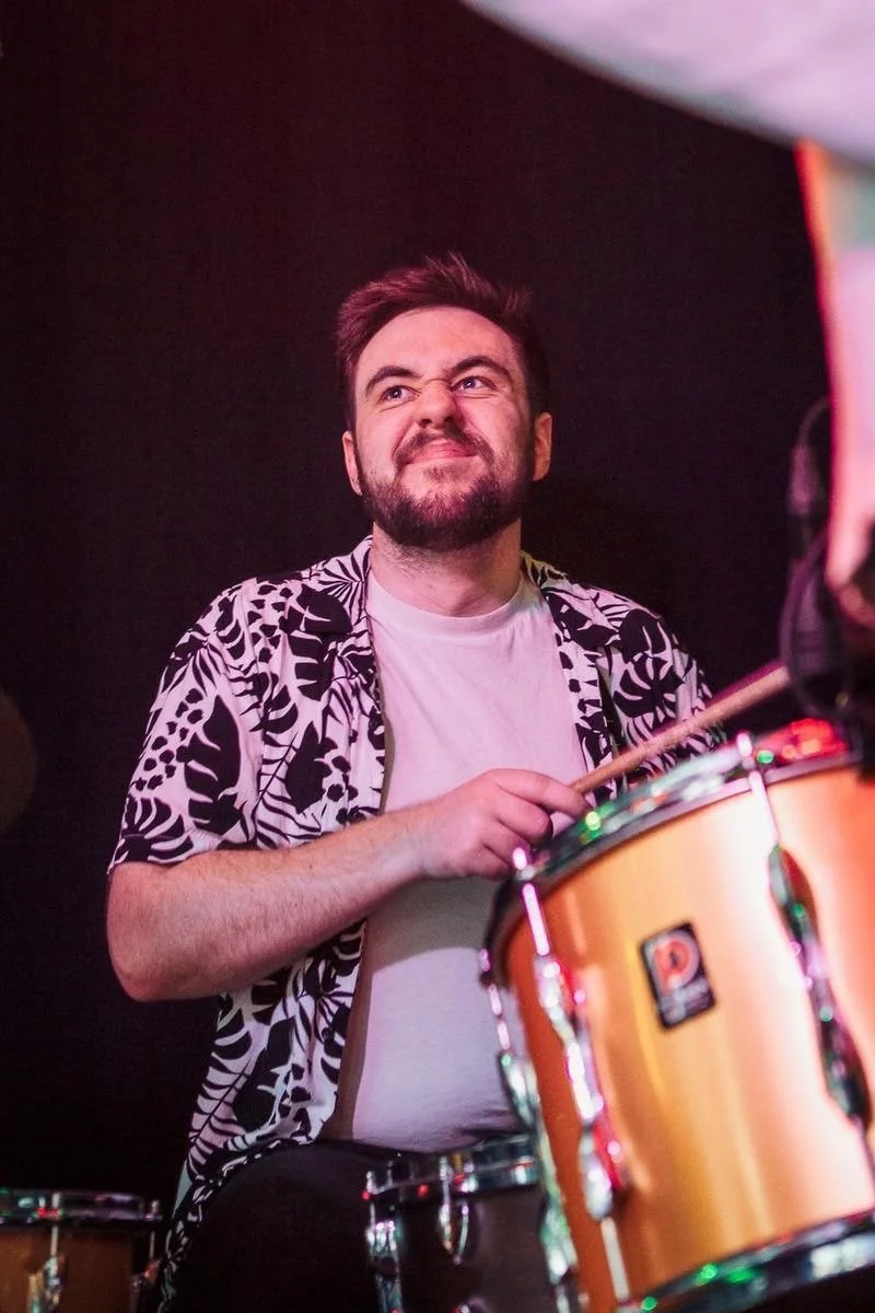 A man with a beard making a face while playing drums on stage, wearing a patterned shirt over a white t-shirt, with colorful stage lighting.