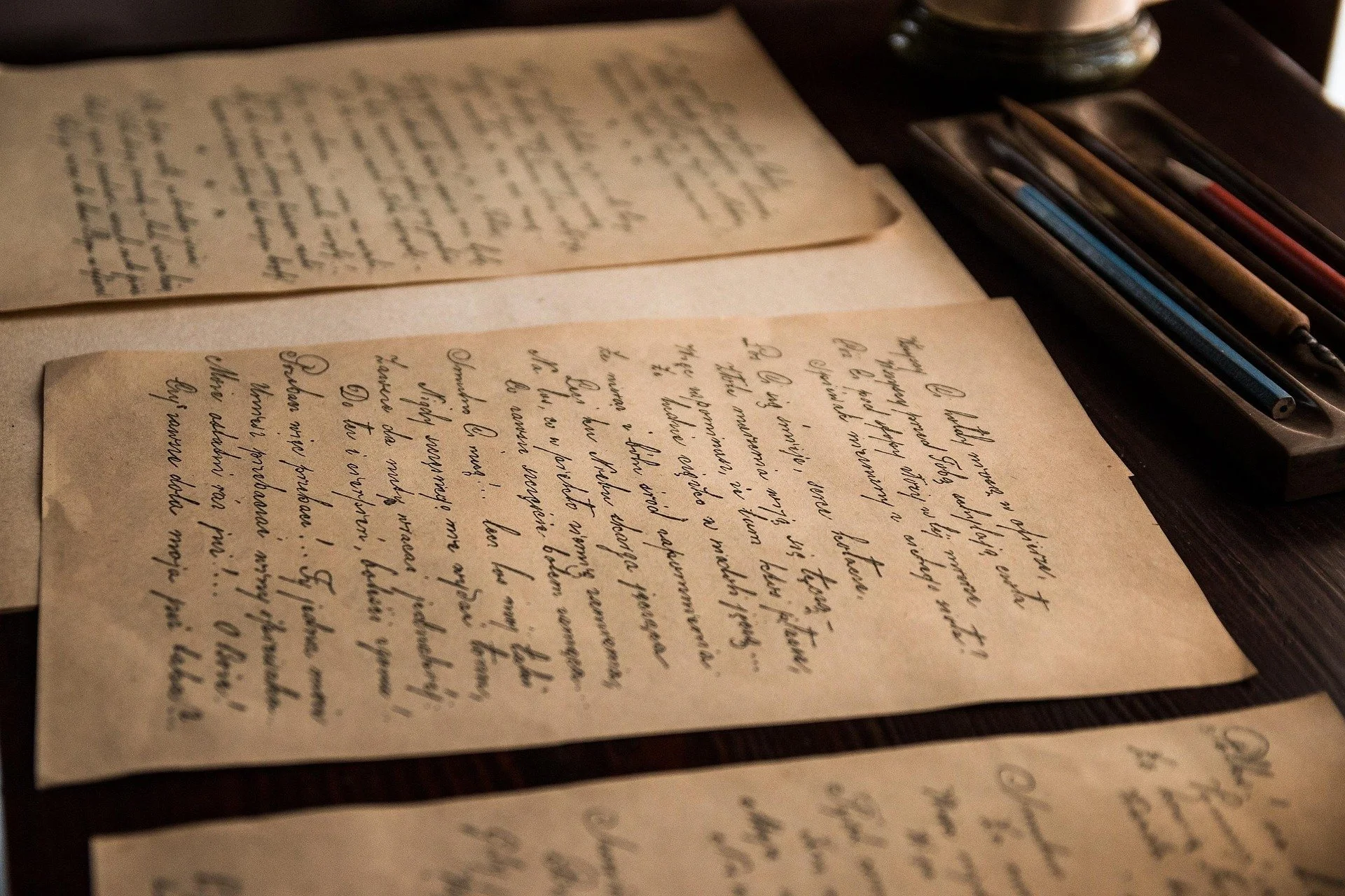 Handwritten letters on yellowed paper, placed on a dark wooden table, with a container holding pens in the background.