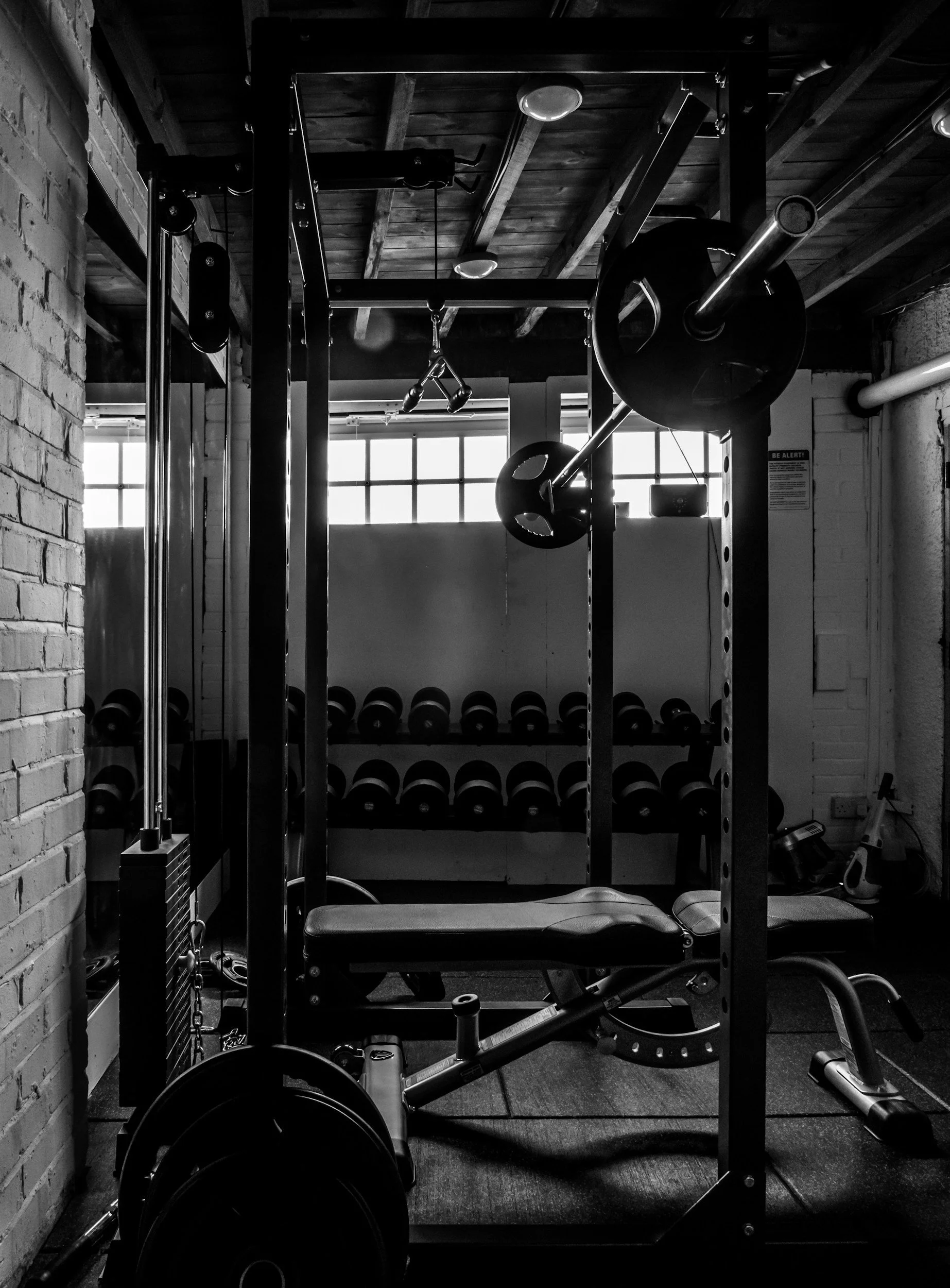 Black and white photo of a gym with weightlifting equipment, including a barbell with weights on a squat rack in the foreground, and a row of dumbbells on a rack in the background, with a brick wall on the left and large windows letting in natural light.