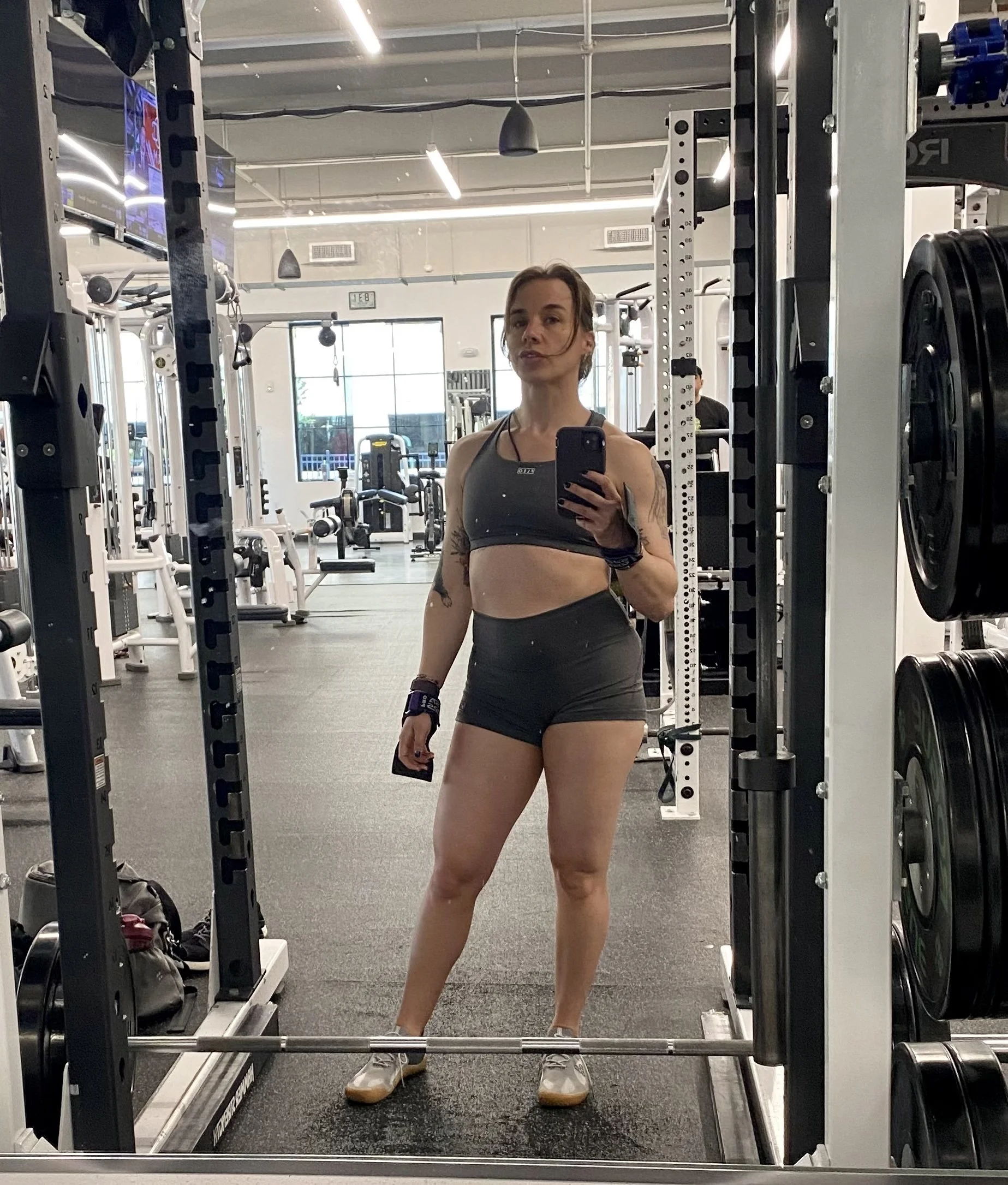 A woman taking a mirror selfie at the gym, wearing workout clothes and sneakers, with gym equipment visible in the background.