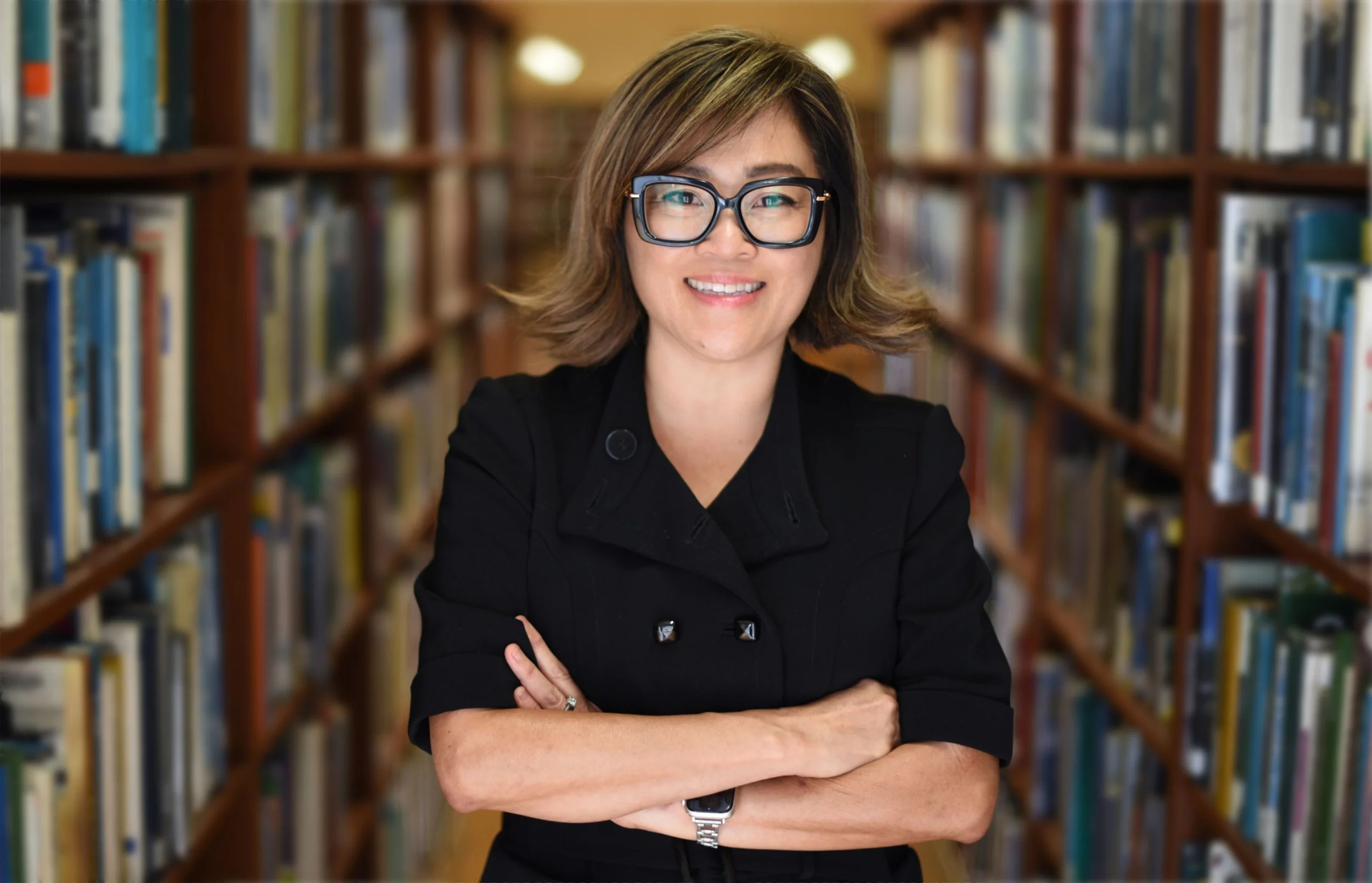 Woman with short brown hair, black glasses, and a black blazer standing in a library with arms crossed, smiling.
