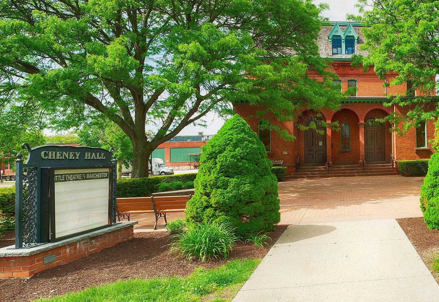 A picture of the entrance to Cheney Hall in Manchester Connecticut. A brick building with arched windows and a small tower, surrounded by trees and shrubs, with a sidewalk leading to the entrance.