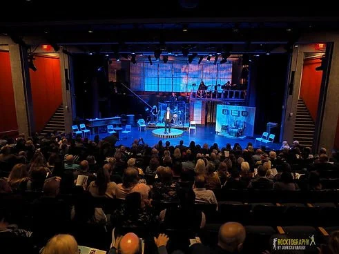 Interior of the Bell Theater in Holmdel, New Jersey. Audience watching a live performance or presentation on a stage with a presenter, set with lights, decorative background, and various props.