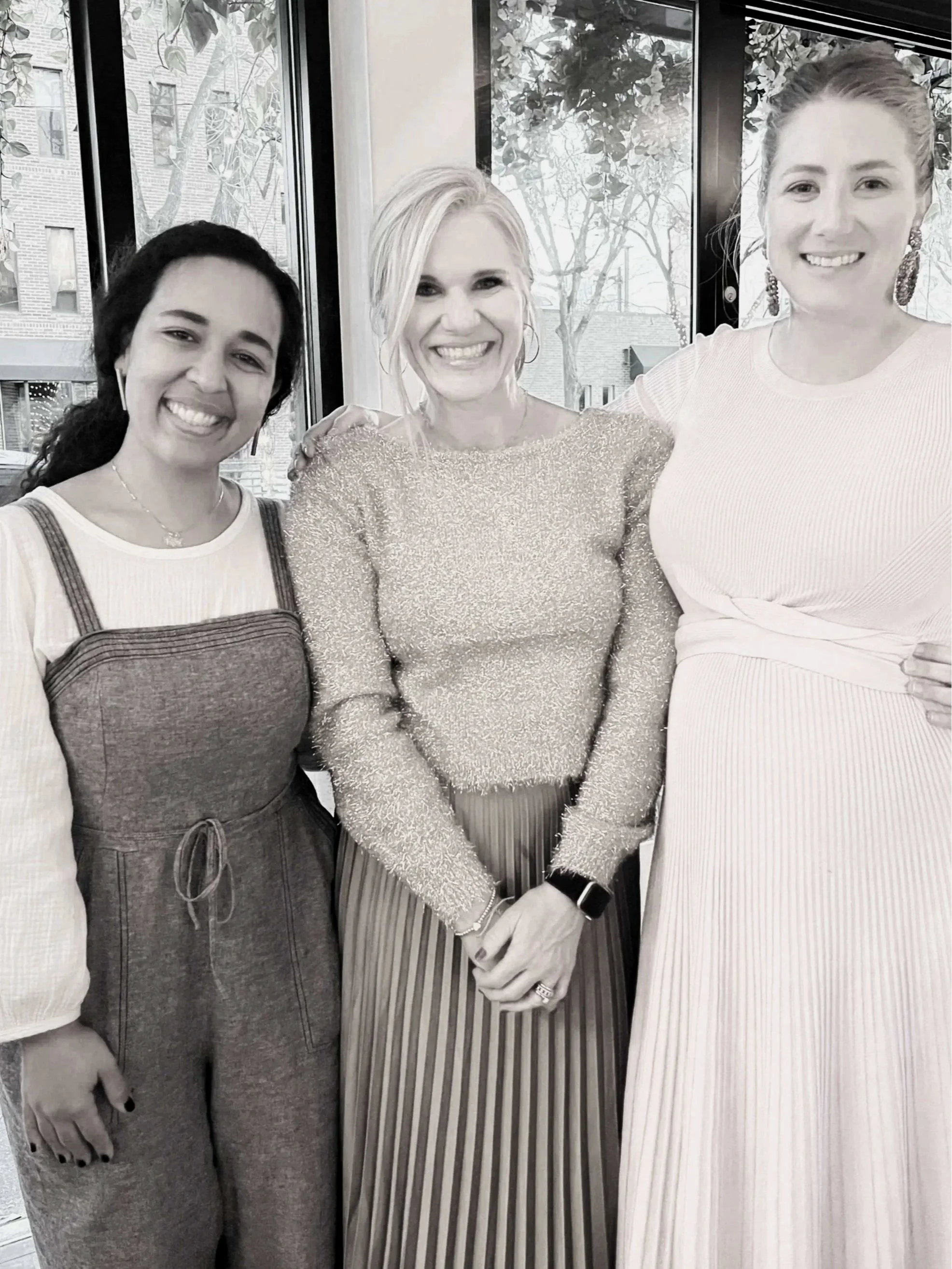 Three women are standing together and smiling in an indoor setting near large windows with trees and buildings visible outside.
