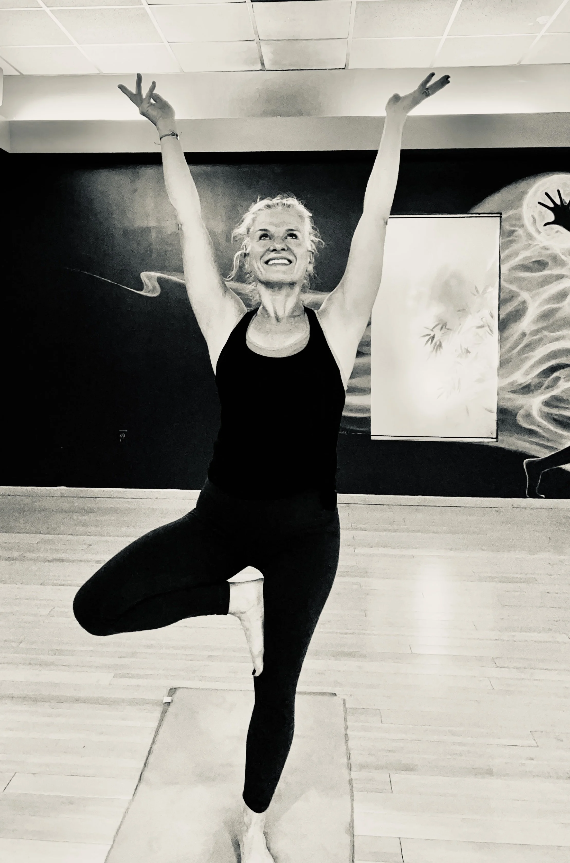 A woman practicing yoga on a mat in a studio, standing on one leg with the other foot resting on her inner thigh, arms raised above her head in a tree pose.