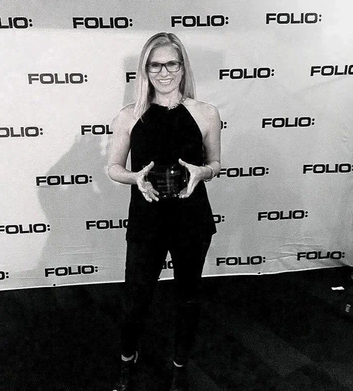 Woman standing in front of a backdrop with the logo 'FOLIO:' repeatedly printed, holding an award or trophy, smiling.