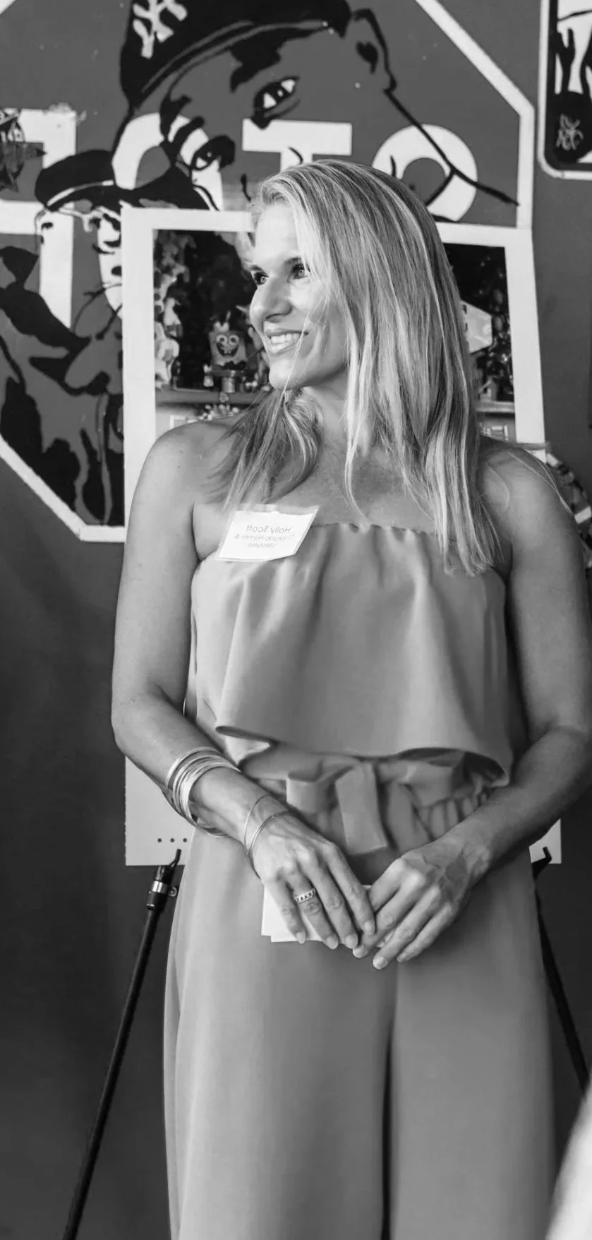 A smiling woman with long blonde hair standing in front of a colorful wall mural, wearing a strapless top, wide-legged pants with a tie at the waist, and jewelry, holding her hands together.