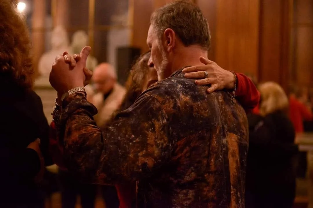 An older man and woman are dancing together in a warm, wooden-paneled room with other people in the background.