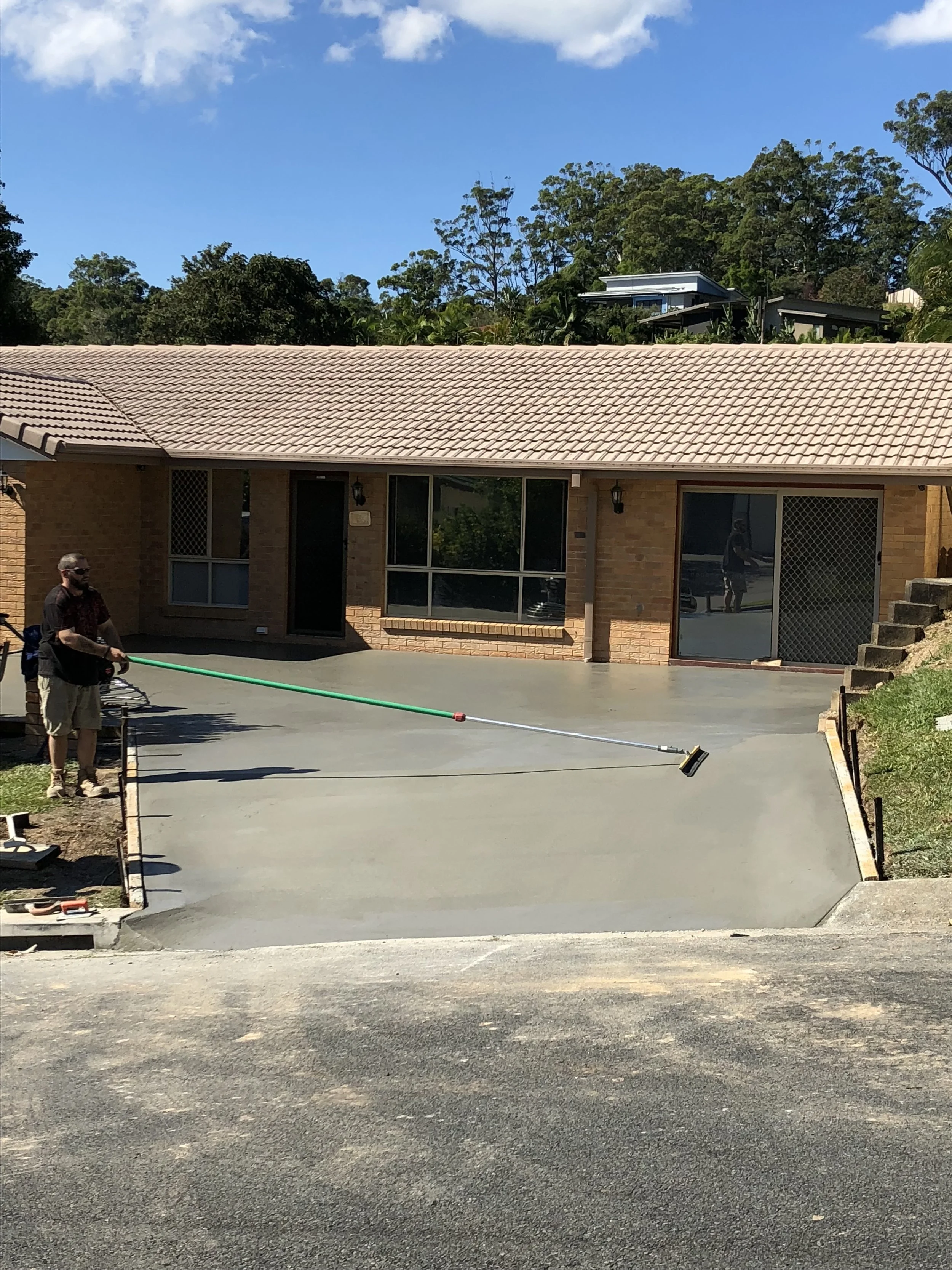 Man smoothing freshly poured concrete on a driveway in front of a brick house with large windows and a sliding glass door.