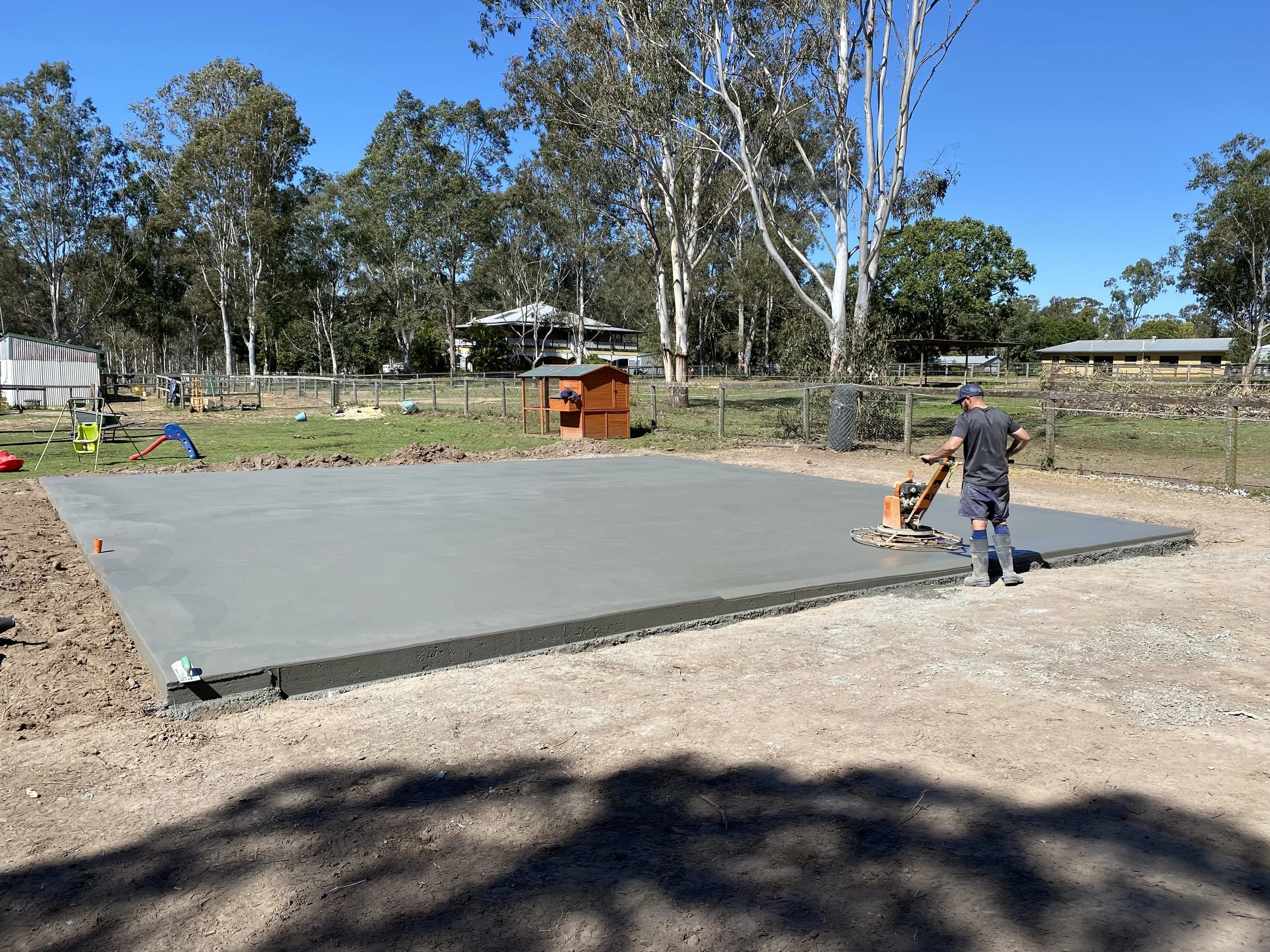 A person is working on a freshly poured concrete slab in a backyard with trees, a fence, and some play equipment in the background.