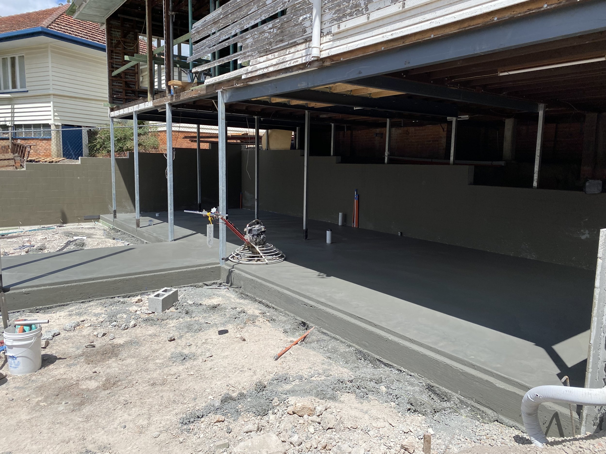 Construction site with freshly poured concrete slab, metal support posts, and partially built wooden structure over a house.