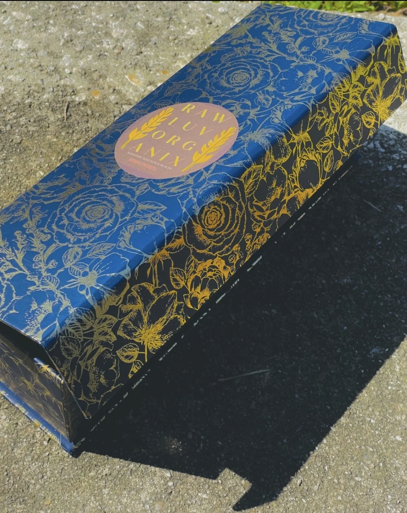 A rectangular box with a dark blue background and gold floral patterns. The top features a pink circle with gold text that says 'Raw Aliv' and 'Luxury'. The box is placed outdoors on a paved surface.