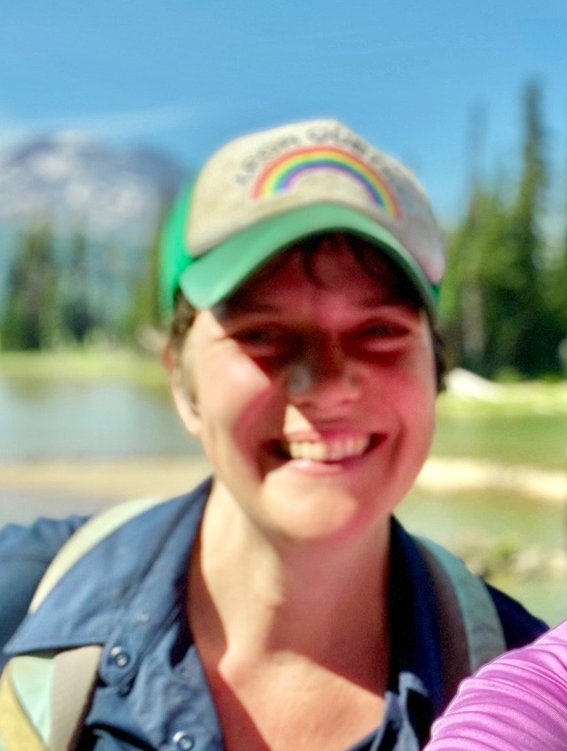 A smiling woman wearing a baseball cap with a rainbow design, outdoors near a body of water, with greenery and trees in the background.