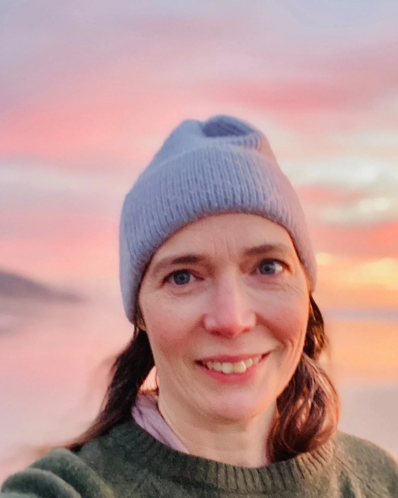 A smiling woman wearing a gray knit beanie and green sweater outdoors during sunset with pink and orange sky in background.