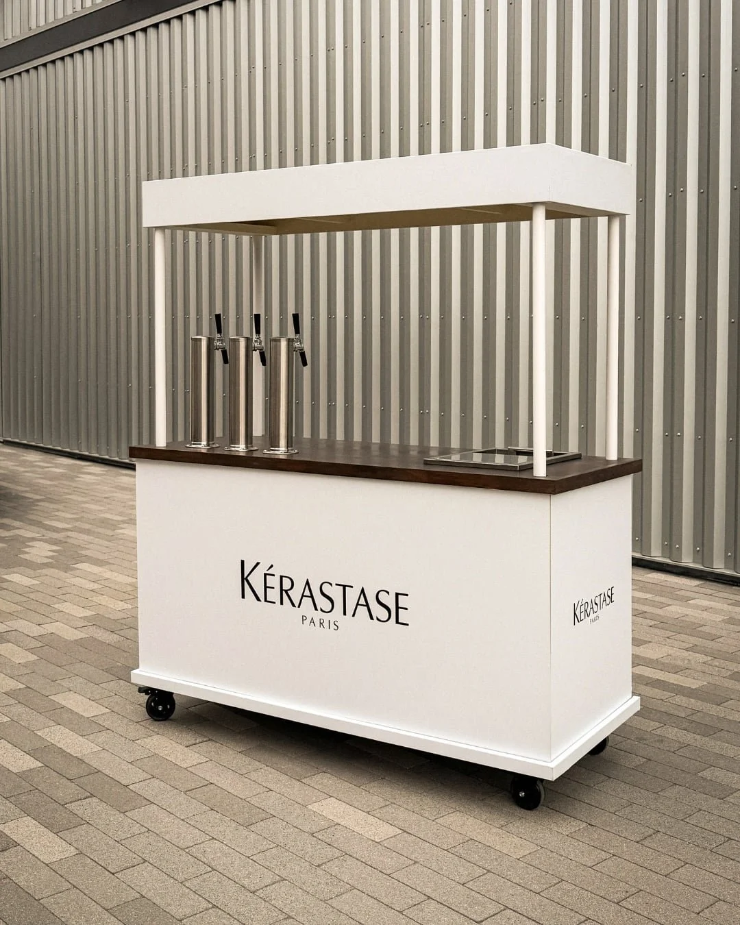 A mobile kiosk cart with a white front panel and black wheels, branded with 'Kérastase Paris', set against a metal wall background. The cart has a dark wooden countertop, three silver beverage dispensers with black handles, and a small flat panel on the right.