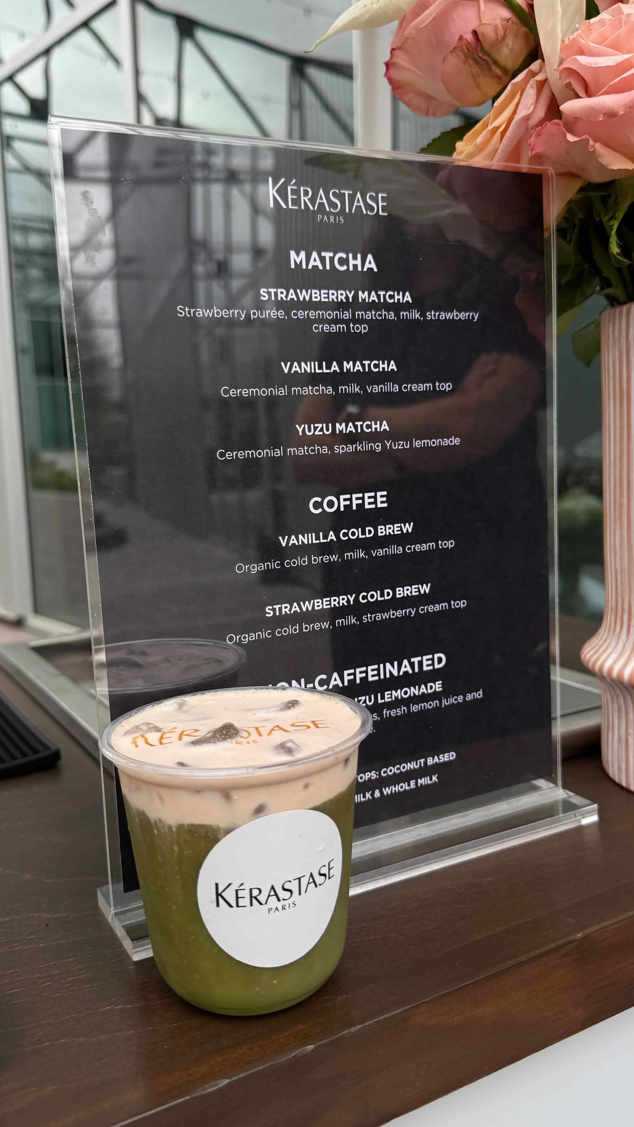 A green matcha latte with ice and a layer of foam in a clear cup, placed on a wooden counter, with a menu behind it listing various matcha and coffee drinks.