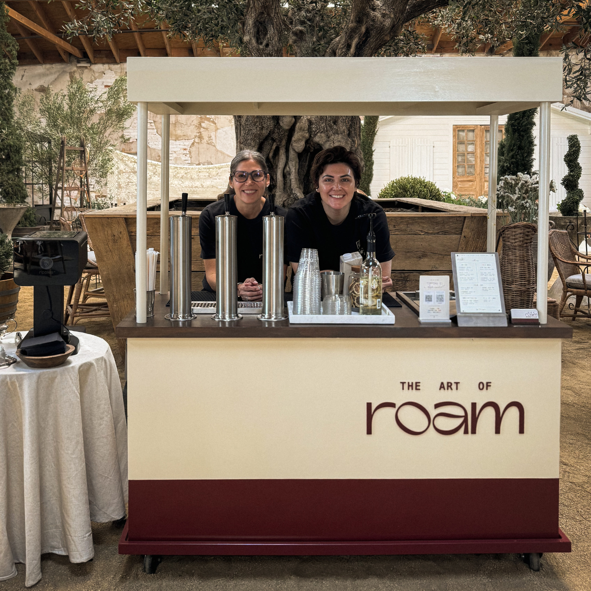 Two women behind a coffee stand with the sign 'The Art of Roam' in a rustic, outdoor setting with trees and plants.