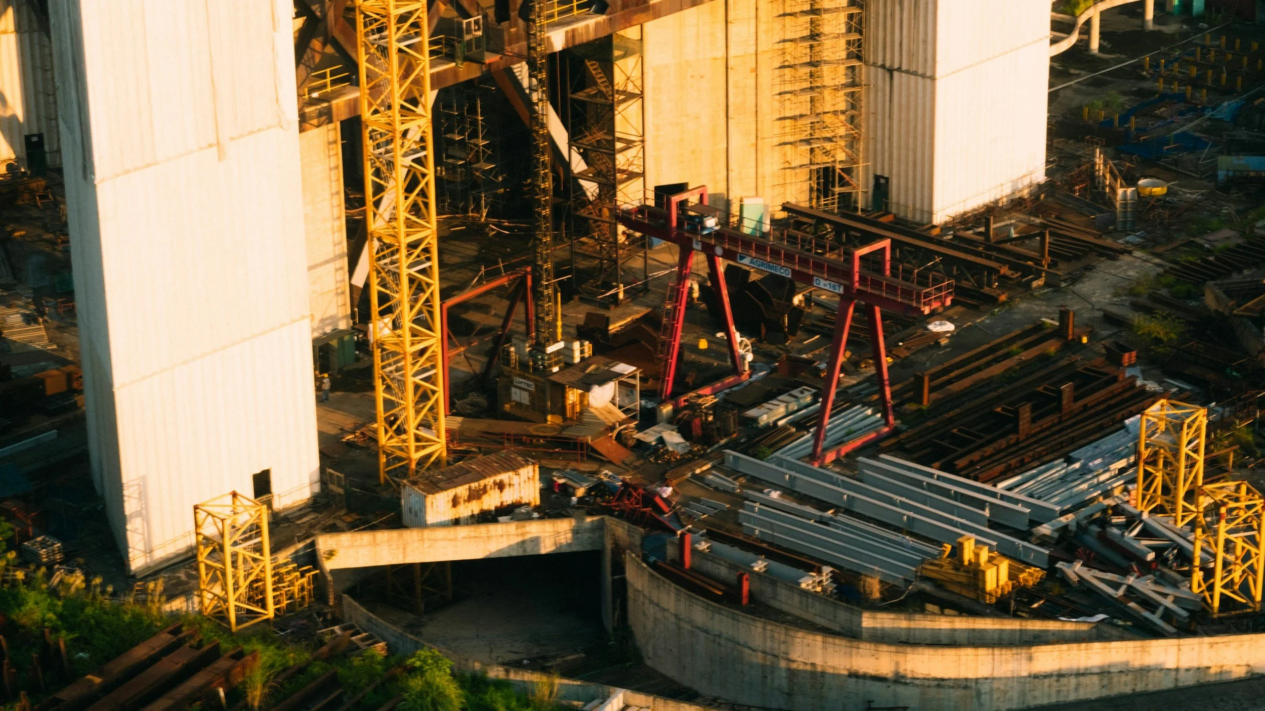 An aerial view of a construction site with large yellow cranes, stacked steel beams, and construction materials, with a partially built structure and surrounding urban area.