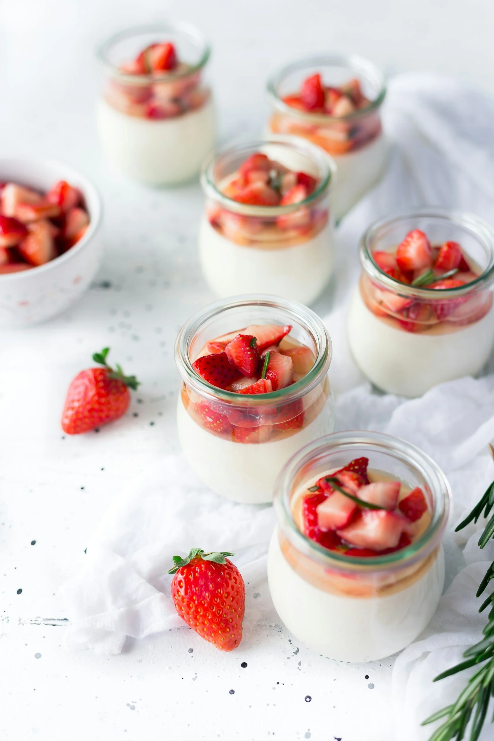 Jars of panna cotta topped with chopped strawberries on a white surface with additional strawberries and greenery.