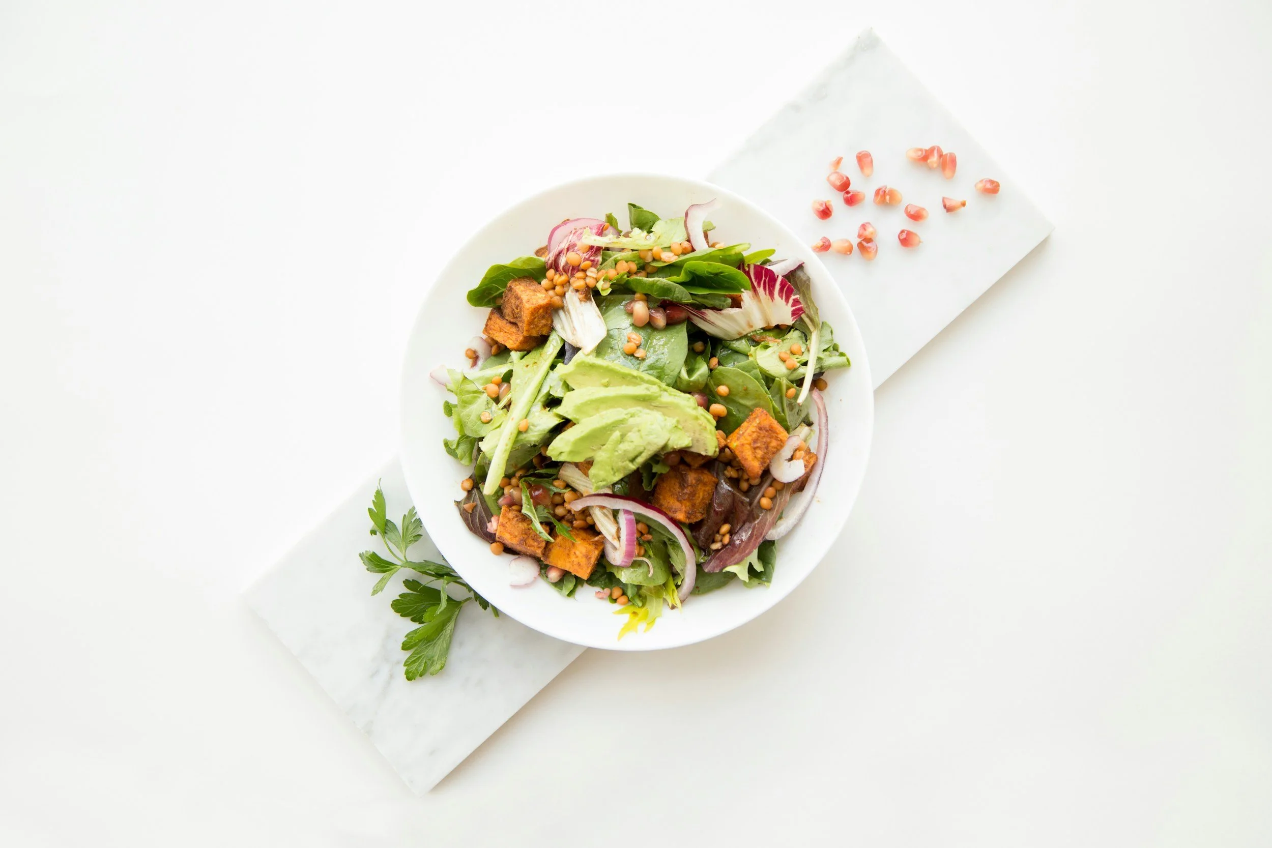 A bowl of fresh green salad with avocado slices, mixed greens, red onions, grilled tofu, and sunflower seeds, placed on a white marble slab with pomegranate seeds on a napkin nearby and a sprig of parsley.