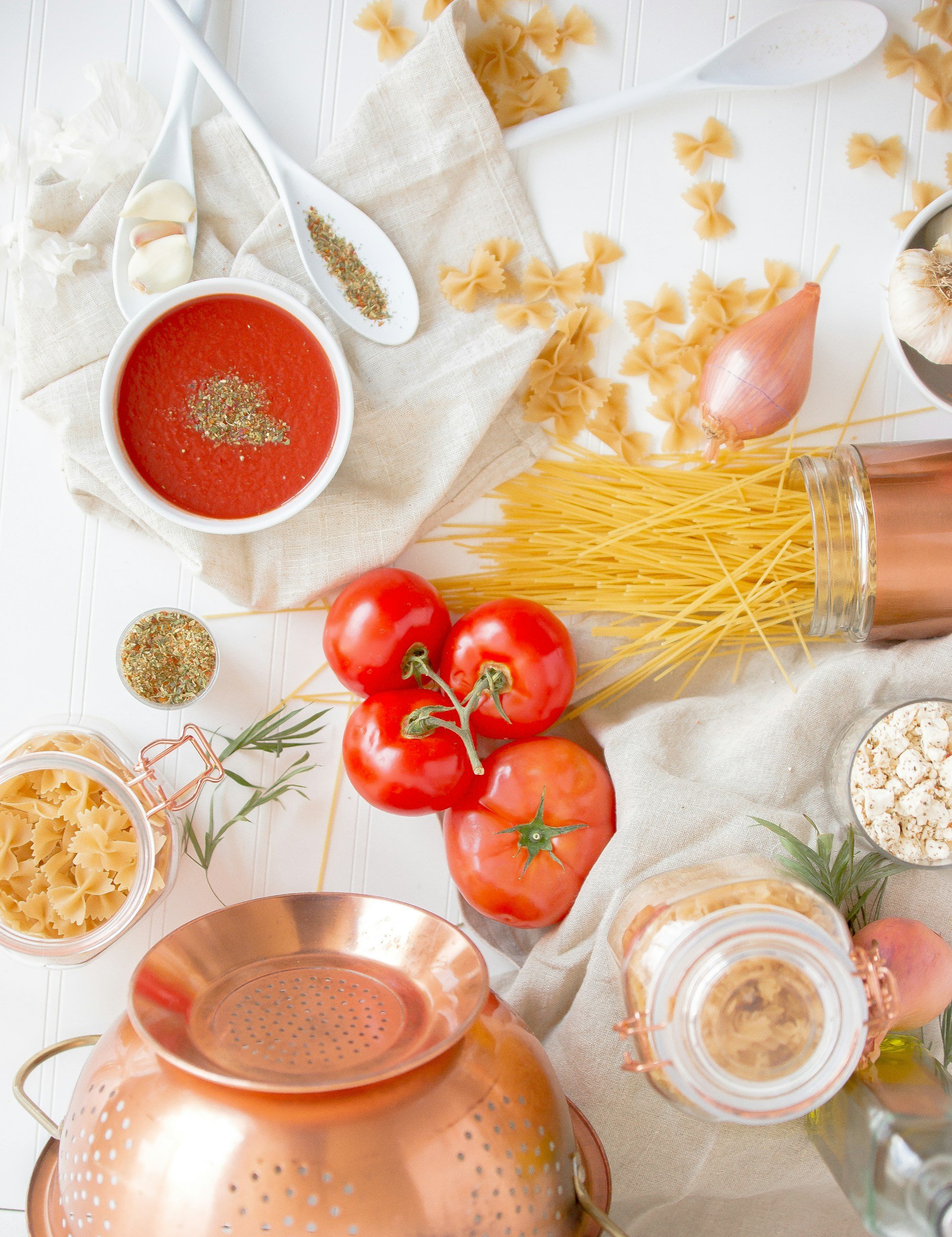 Kitchen countertop with tomatoes, pasta, garlic, onion, tomato sauce, spices, and cooking utensils for preparing spaghetti.