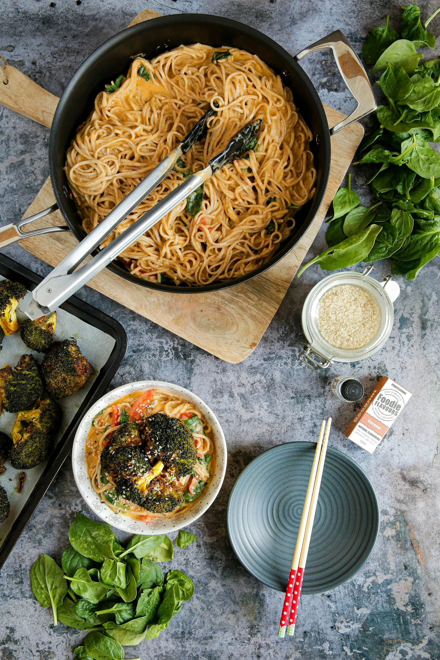 A top-down view of a table with a pot of cooked ramen noodles, a bowl of ready-to-eat ramen with vegetables, roasted broccoli, a small pile of fresh spinach leaves, a small jar and a box of flavoring, chopsticks on a blue plate, and a pair of tongs resting on the pot.