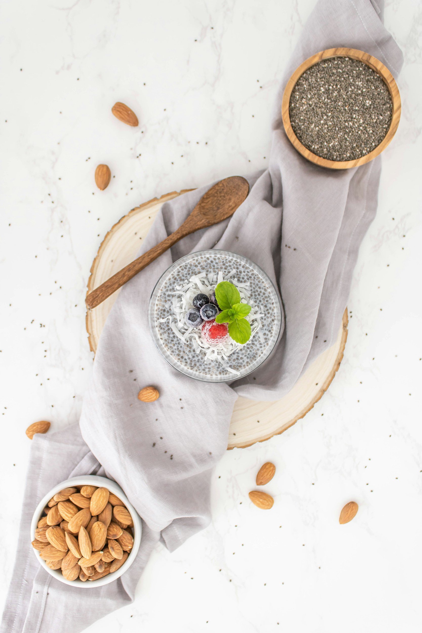 A chia seed pudding topped with blueberries, a raspberry, and mint leaves, served in a glass bowl, surrounded by almonds, on a wooden board with a grey cloth, chia seeds in a wooden bowl, and a small wooden spoon.