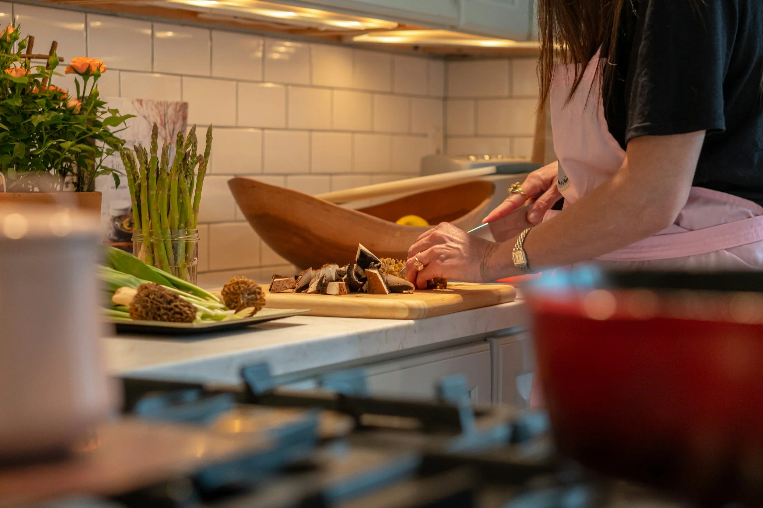 Person chopping mushrooms on a wooden cutting board in a kitchen with plants and various kitchen items around.