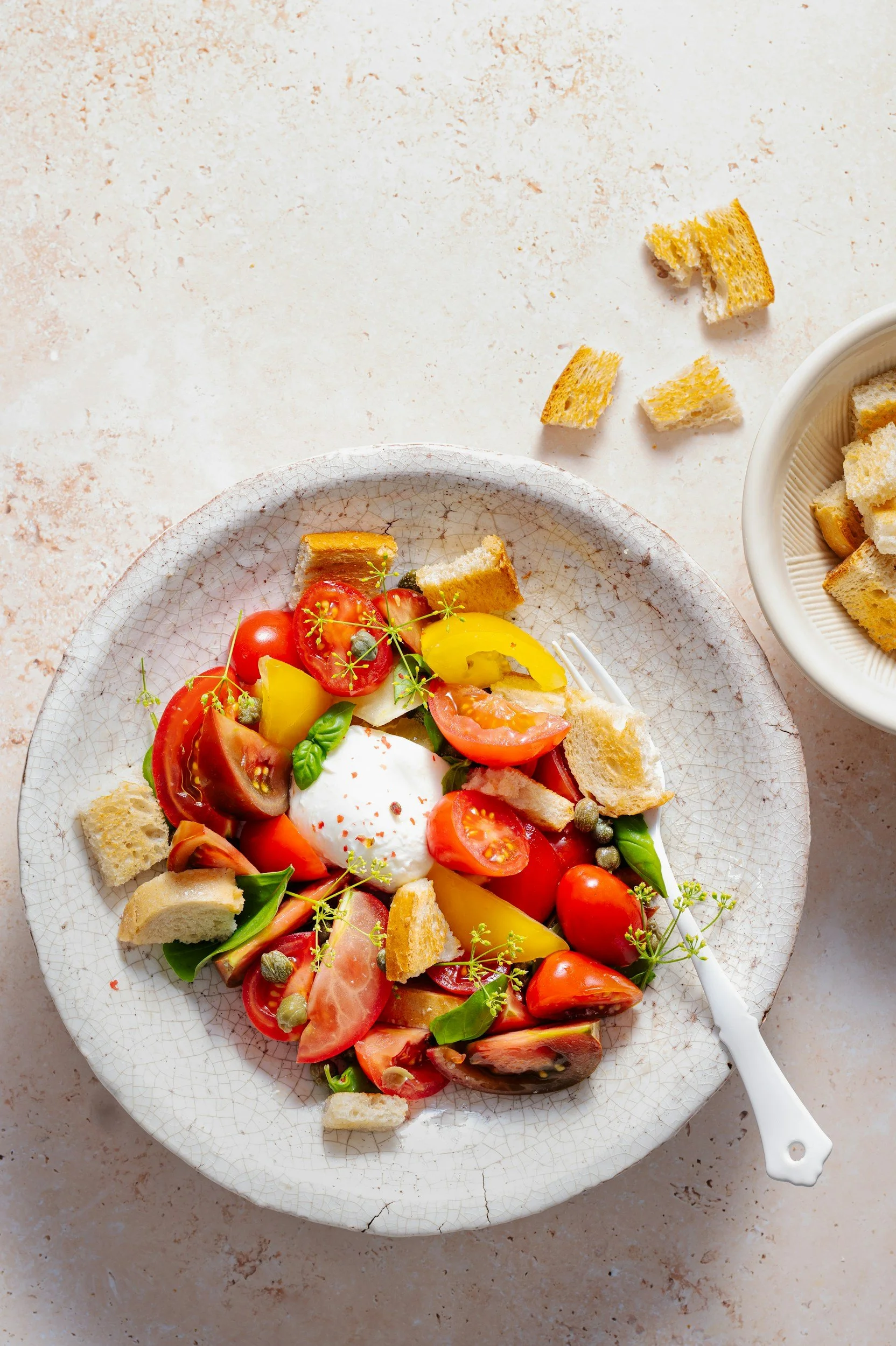 A plate of fresh tomato salad with torn bread pieces, and a poached egg in the center, garnished with herbs, on a light textured surface.