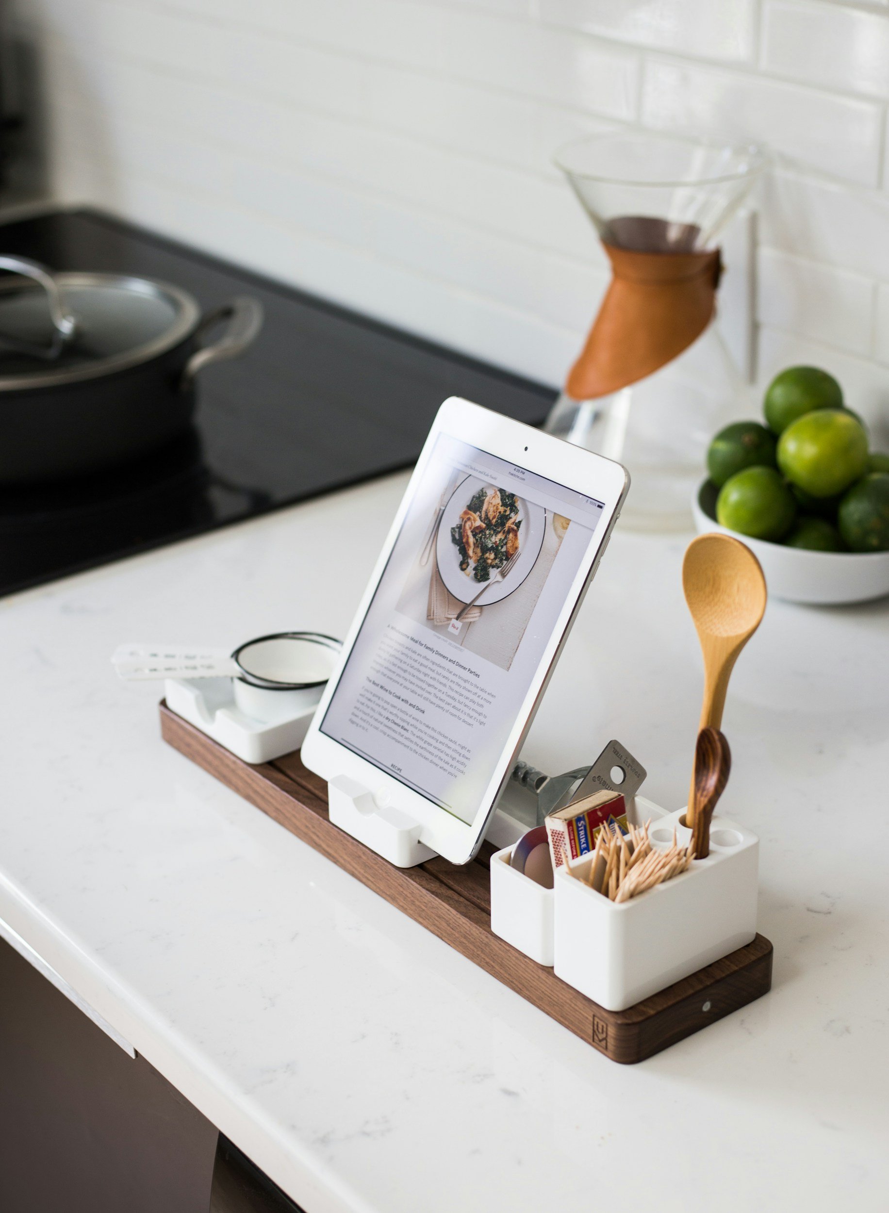A kitchen countertop with a tablet in a stand displaying a recipe, a white container holding wooden spoons and packets, a bowl of limes, and a glass coffee or cocktail with a leather holder, with a stove and black cookware in the background.
