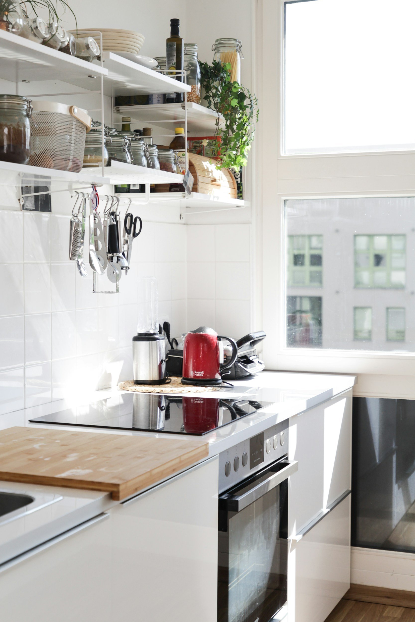 A modern kitchen with a white countertop, a red kettle, a silver toaster, and kitchen utensils hanging on the wall above a white tiled backsplash. There are open shelves with jars, bottles, and plants near large windows letting in natural light.