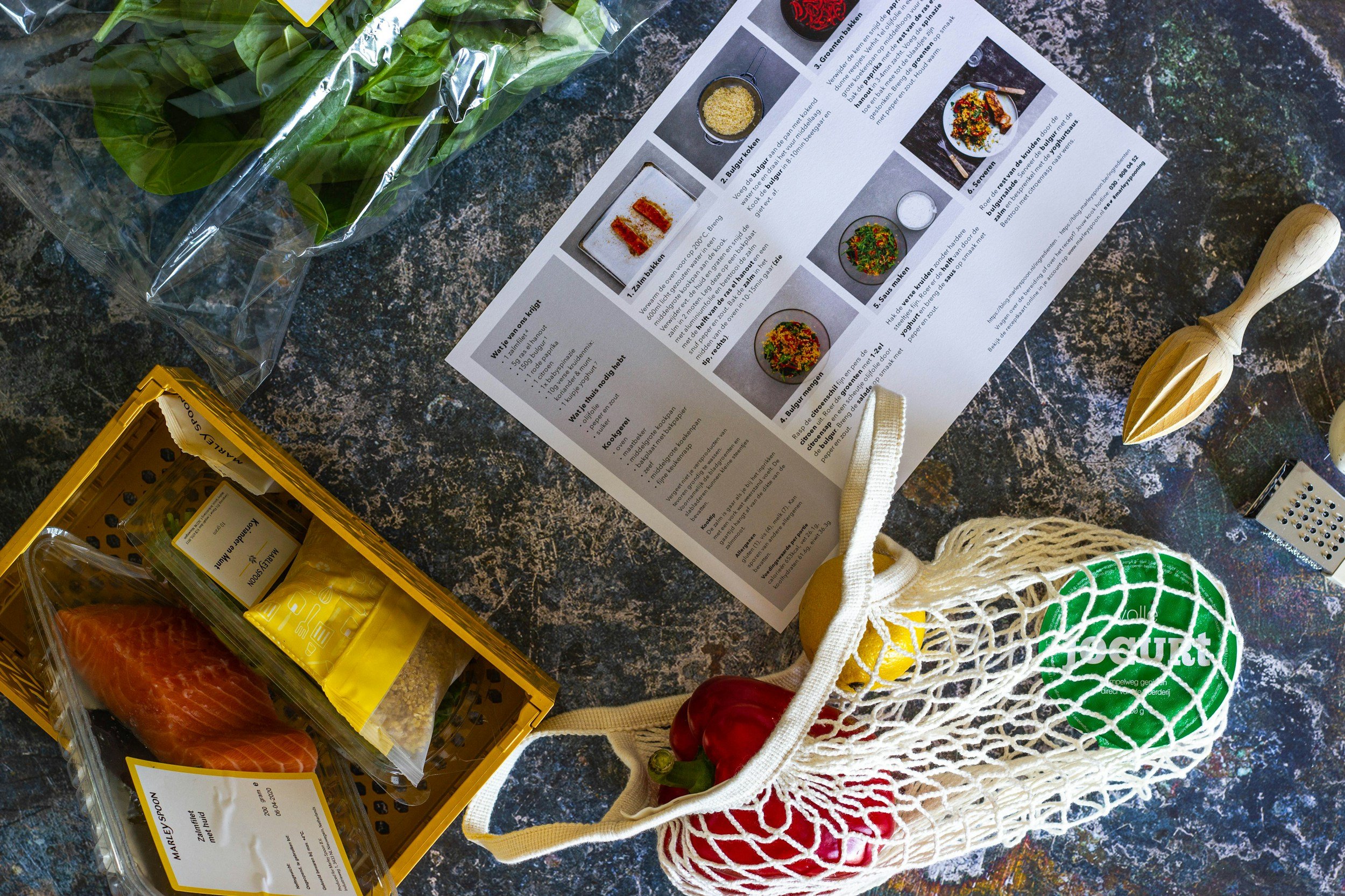 A grocery bag filled with vegetables like red and green peppers, a nearby yellow basket with salmon, spices, and other food items, a scooping tool, and a printed recipe on a dark, textured surface.