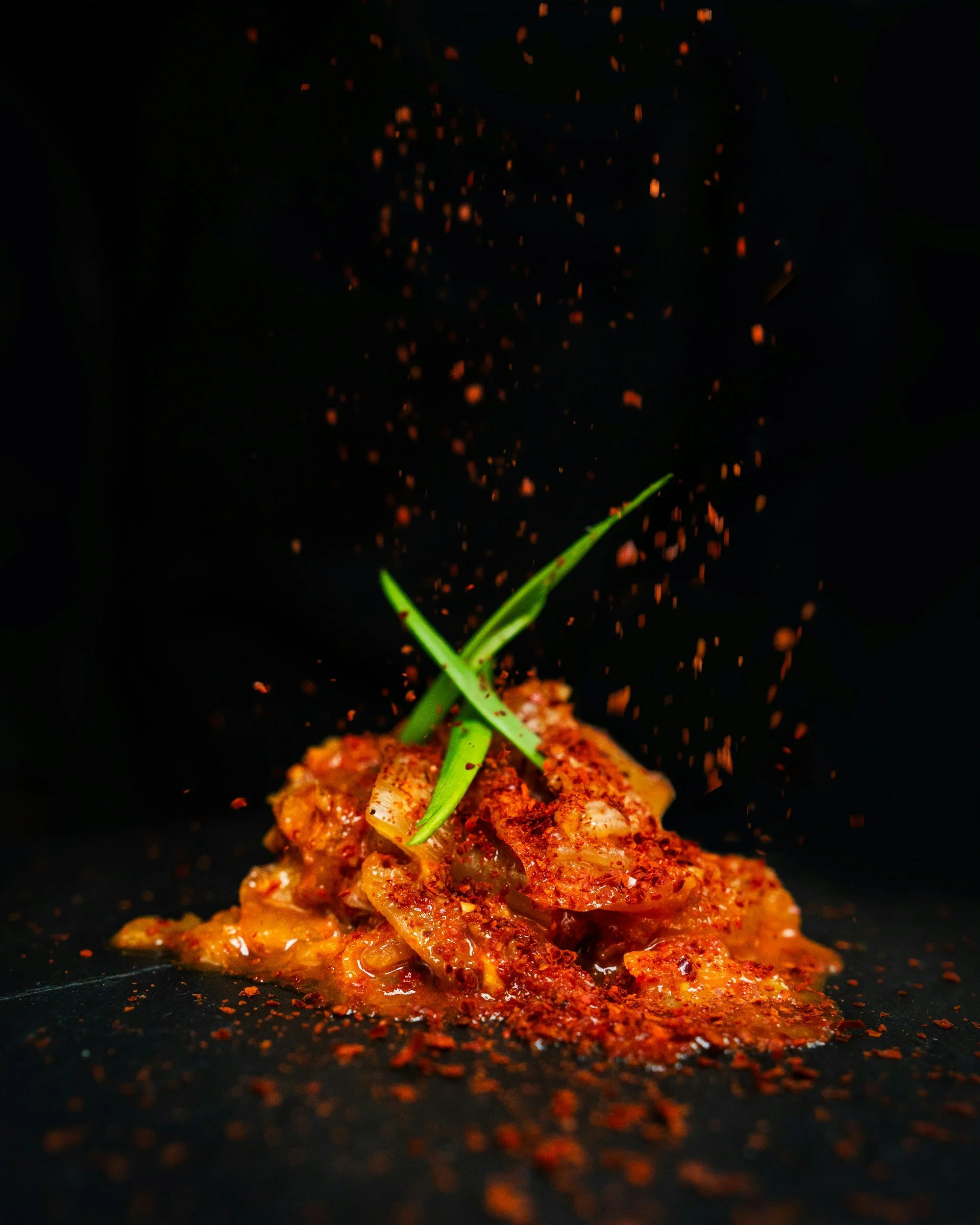Close-up of a chef's knife slicing seasoned meat with chili flakes, green onion garnish, with orange spices and herbs in a dark background.