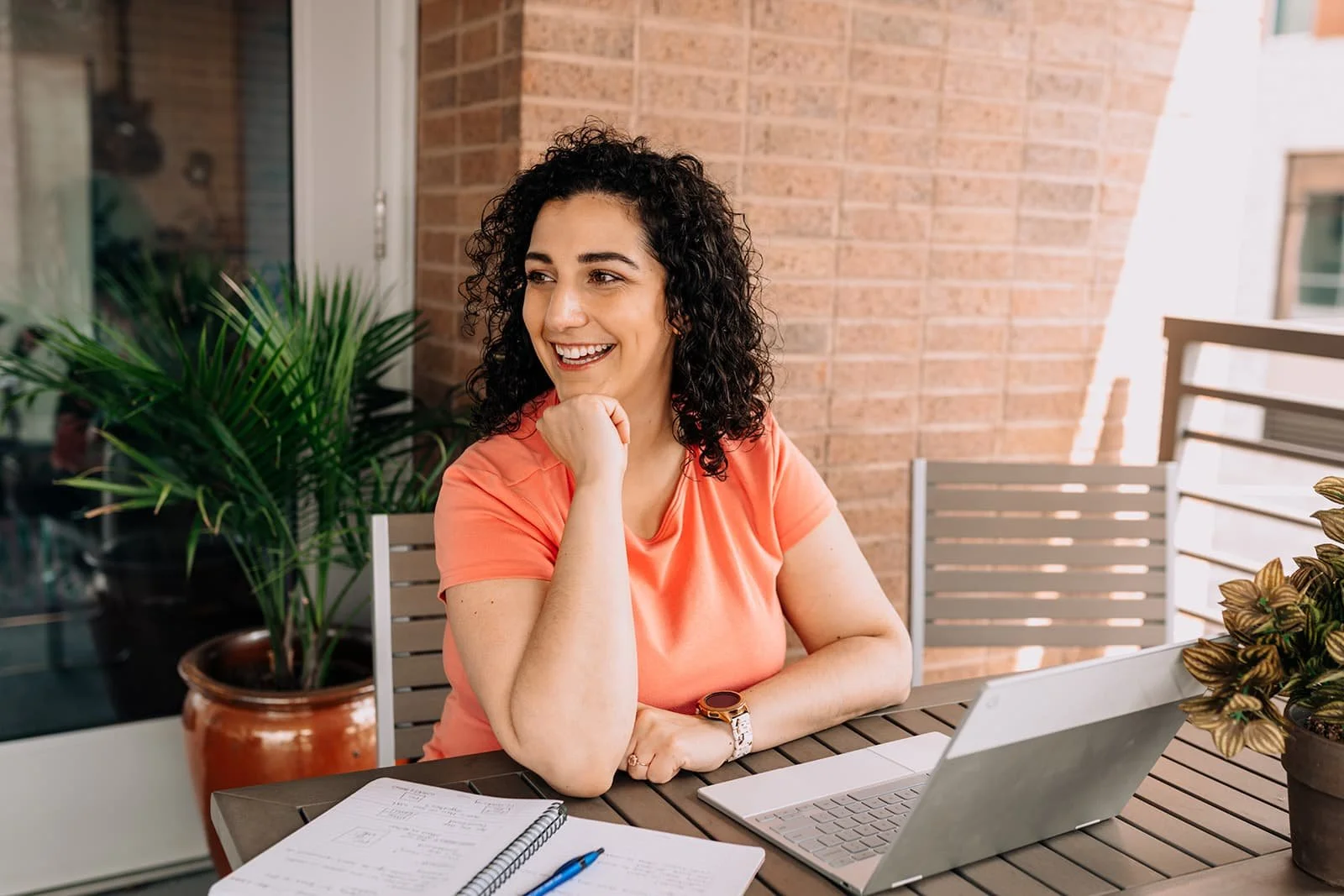 A woman with curly black hair, wearing an orange shirt, sitting at an outdoor table with a laptop, notebook, and pen, smiling.