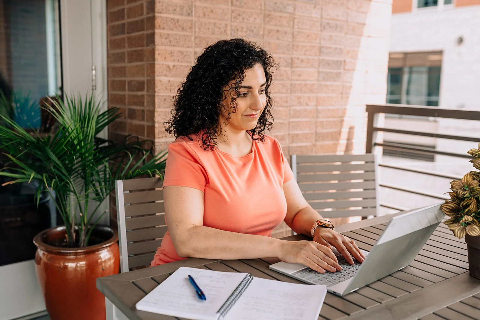 Woman with curly black hair sitting at an outdoor table working on a silver laptop, with a potted plant and a notebook with a pen in front of her.