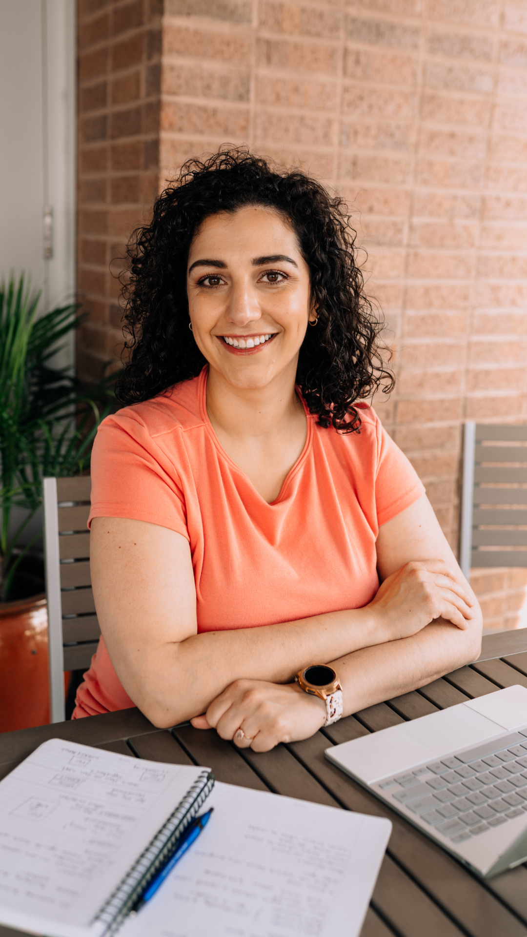 A woman with dark, curly hair smiling and sitting at a table with a notebook, pen, laptop, and documents, in front of a brick wall.