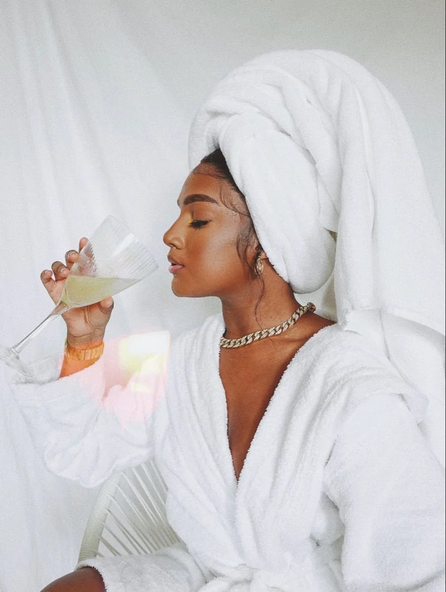 A woman in a white towel and a towel turban is holding a champagne glass with a drink. She is wearing a chain necklace and a watch.