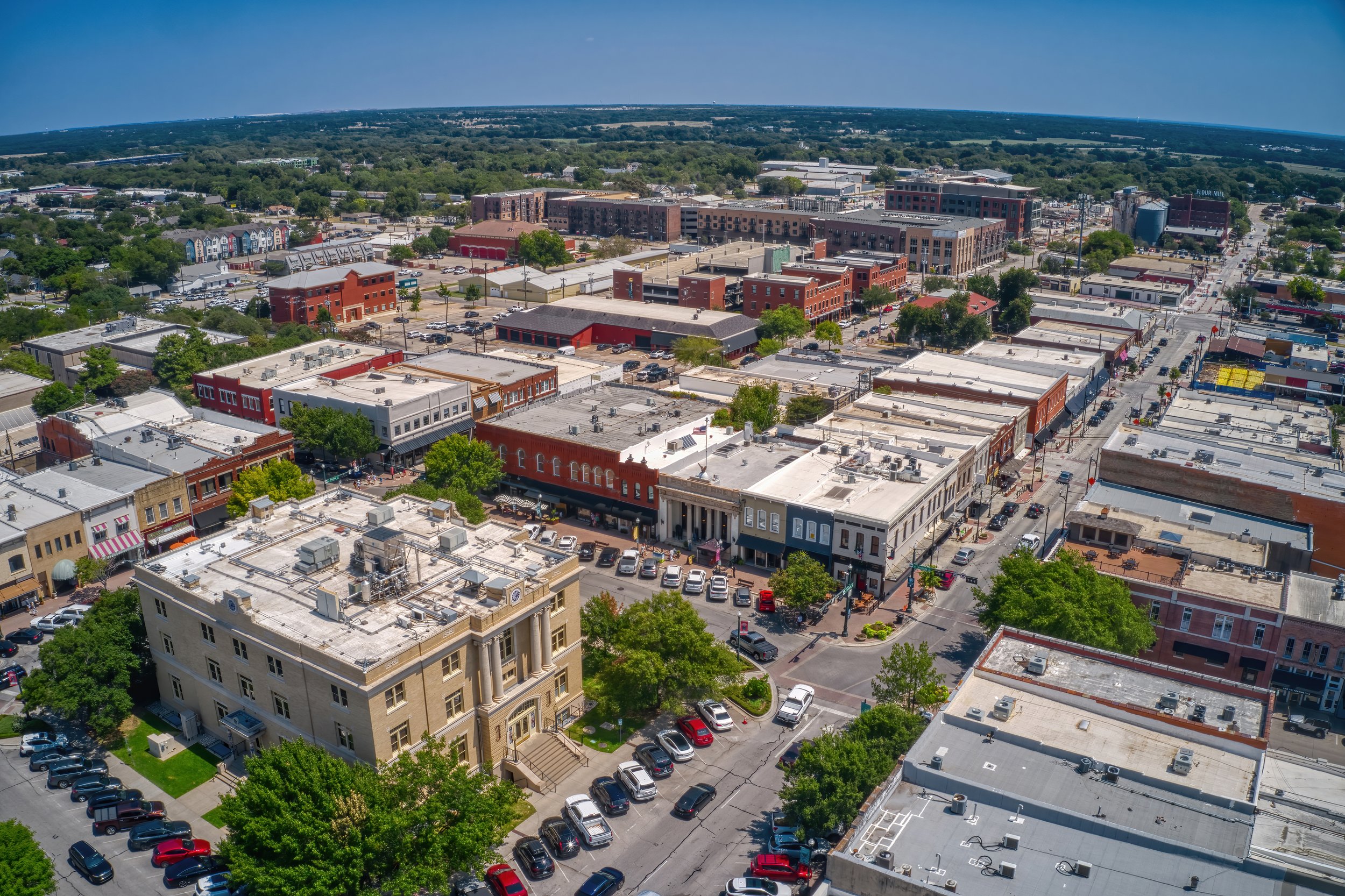 Aerial view of a downtown area with a mix of historic and modern buildings, streets, cars, and green spaces, with trees and clear skies.