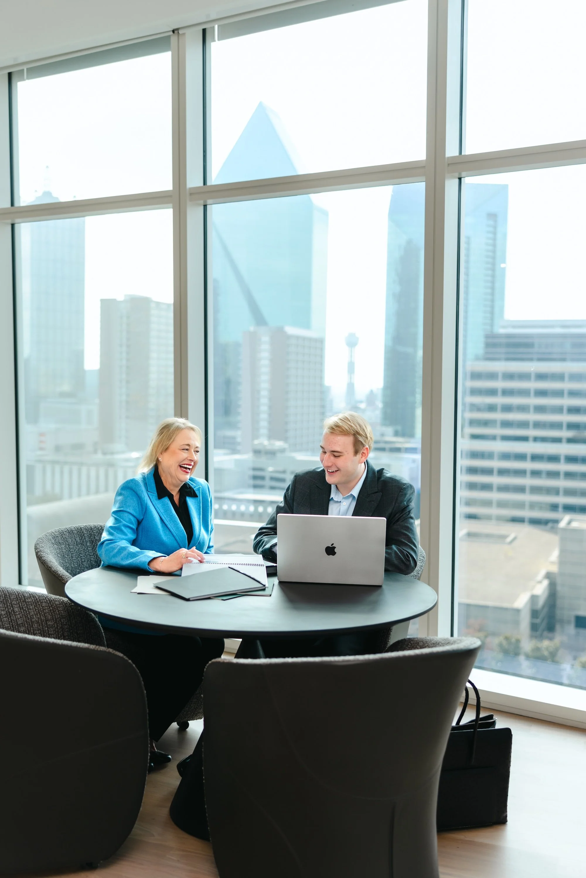 Two business professionals, a woman and a man, sitting at a round table in a high-rise office with large windows showing a city skyline, engaged in a conversation and smiling.