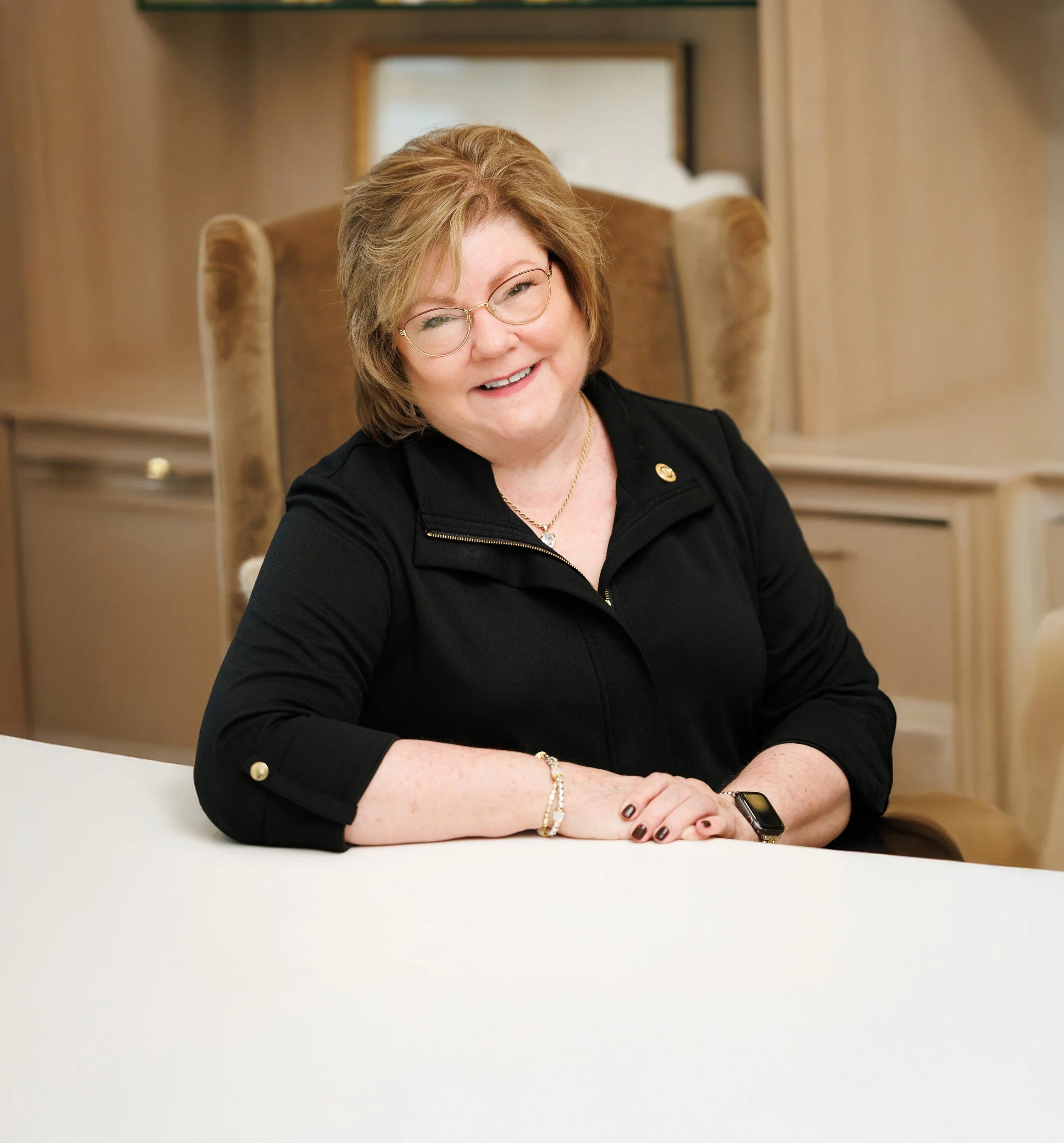 A smiling woman with glasses and short reddish hair sitting at a white table, wearing a black top, gold jewelry, and a smartwatch in a warm-toned room with wooden furnishings.