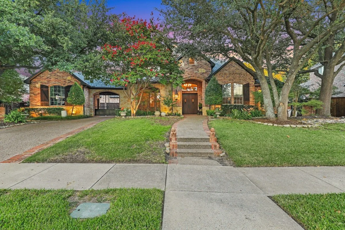 A charming brick house with a lush front yard, a curved pathway leading to the front door, and tall trees providing shade. The house has large windows and an inviting entrance.