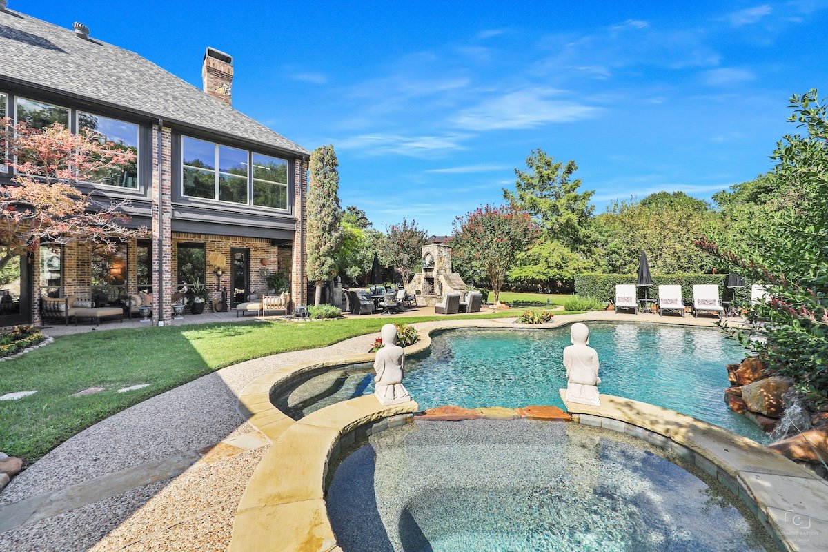 Backyard with a swimming pool, surrounded by green trees and bushes, with patio furniture and a house with large windows in the background.